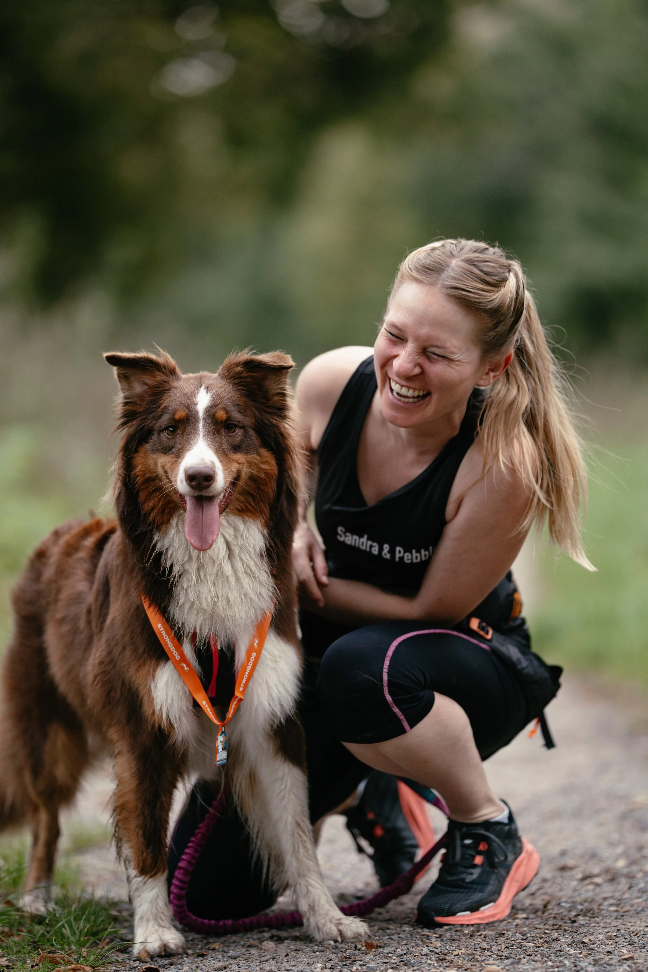 Eine Australien Shepherd Hündin steht neben ihrem lachenden Frauchen.