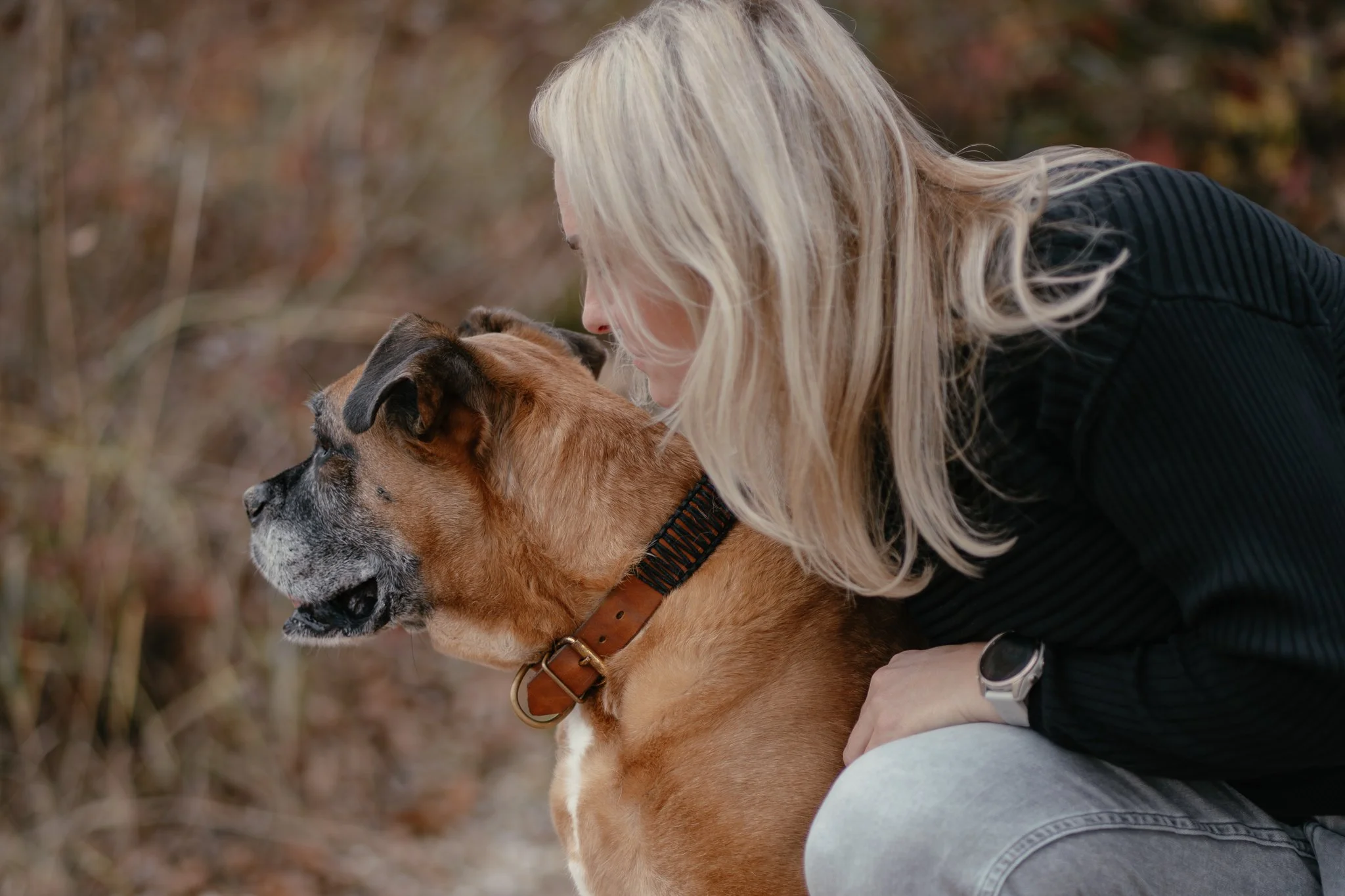 Frauchen umarmt einen braunen Hund, im Hintergrund sind Bäume und herbstliche Blätter.