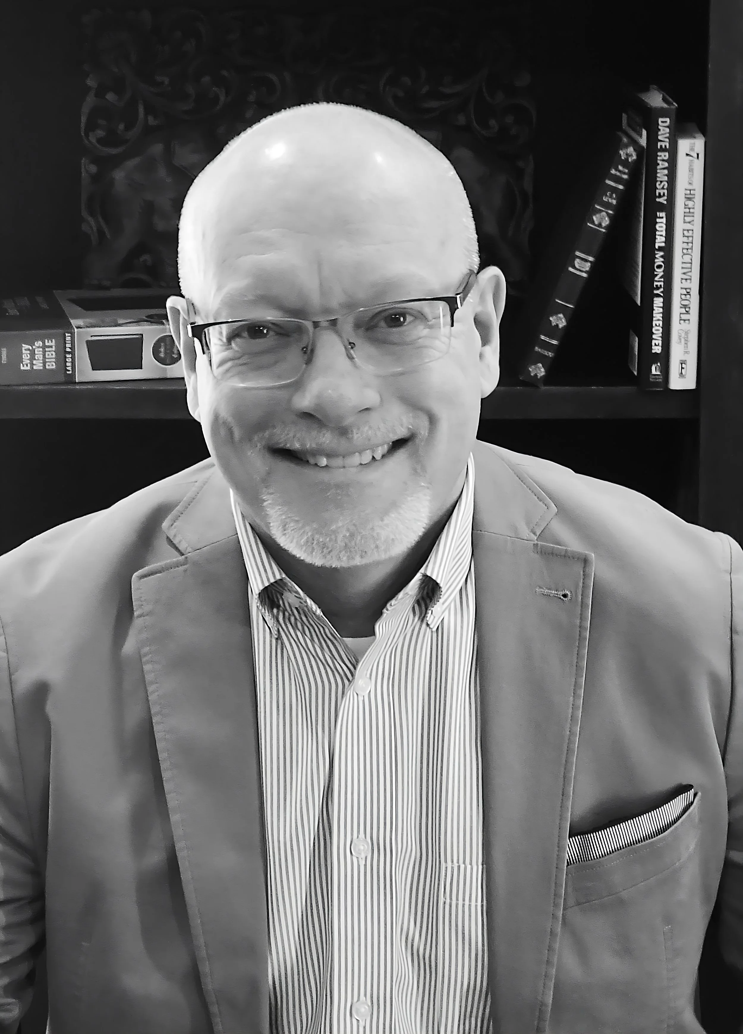A smiling man with glasses, wearing a striped shirt and blazer, sitting in front of a bookshelf with books in the background.