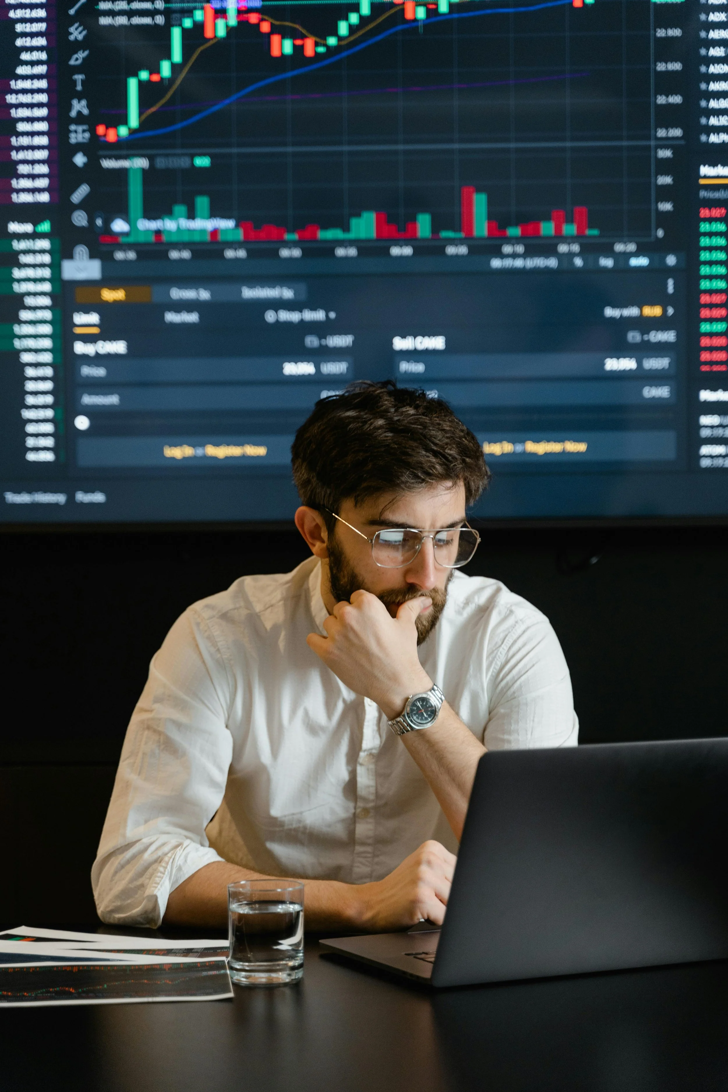 A man in a white shirt with glasses and a wristwatch sitting at a desk, looking at a laptop, with a glass of water and some papers on the desk. Behind him is a large screen displaying a financial trading chart with candlestick patterns, volume bars, and various data.