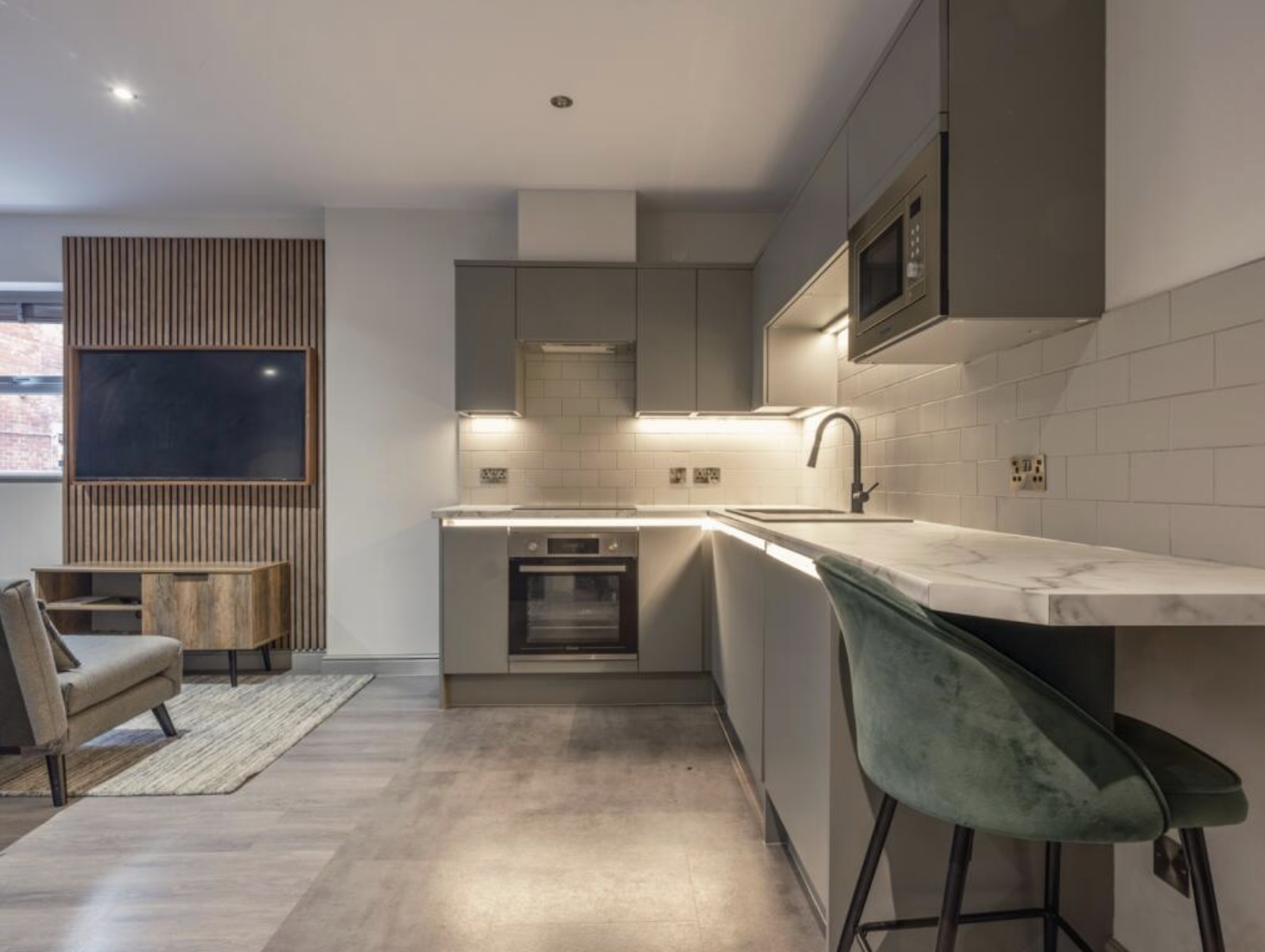 Modern kitchen with gray cabinets, white subway tile backsplash, marble countertop, built-in oven, microwave, and a green velvet barstool. Adjacent to a living area with a TV on a wooden slatted wall and a beige armchair.
