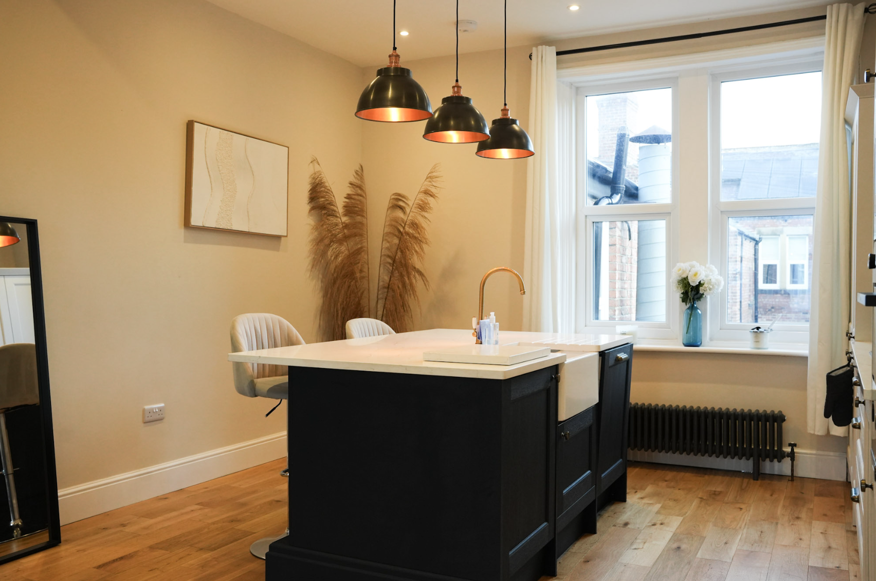 Modern kitchen with black island, beige walls, large window with white curtains, vase of white flowers, pendant lights, and wood flooring.