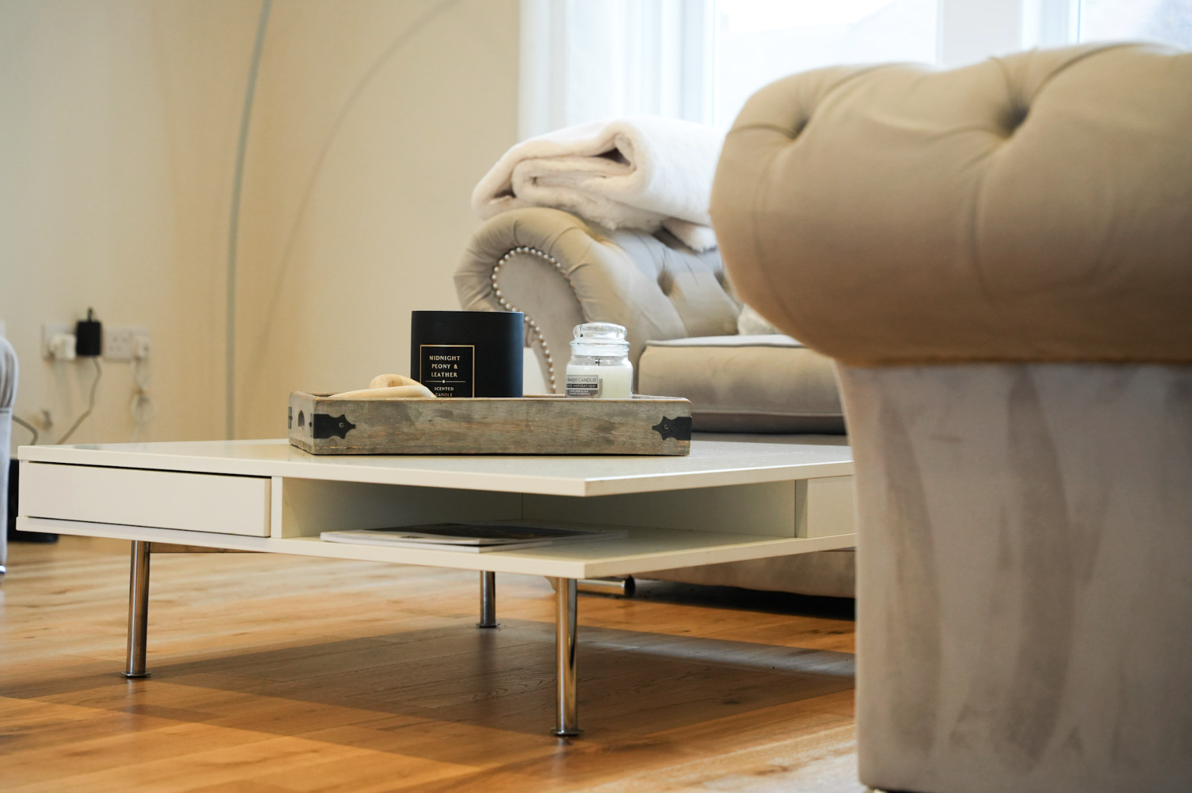Living room with beige sofa, white coffee table with tray holding candle and black container, folded blanket on sofa, hardwood floor, and a window in background.