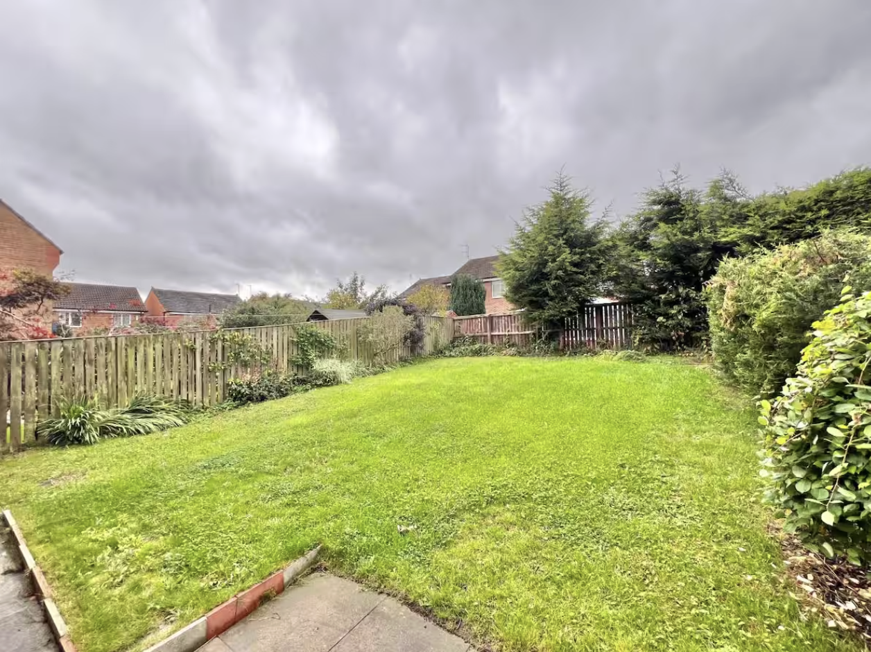 A backyard with a green lawn, surrounded by trees, bushes, and a wooden fence, under a cloudy sky.