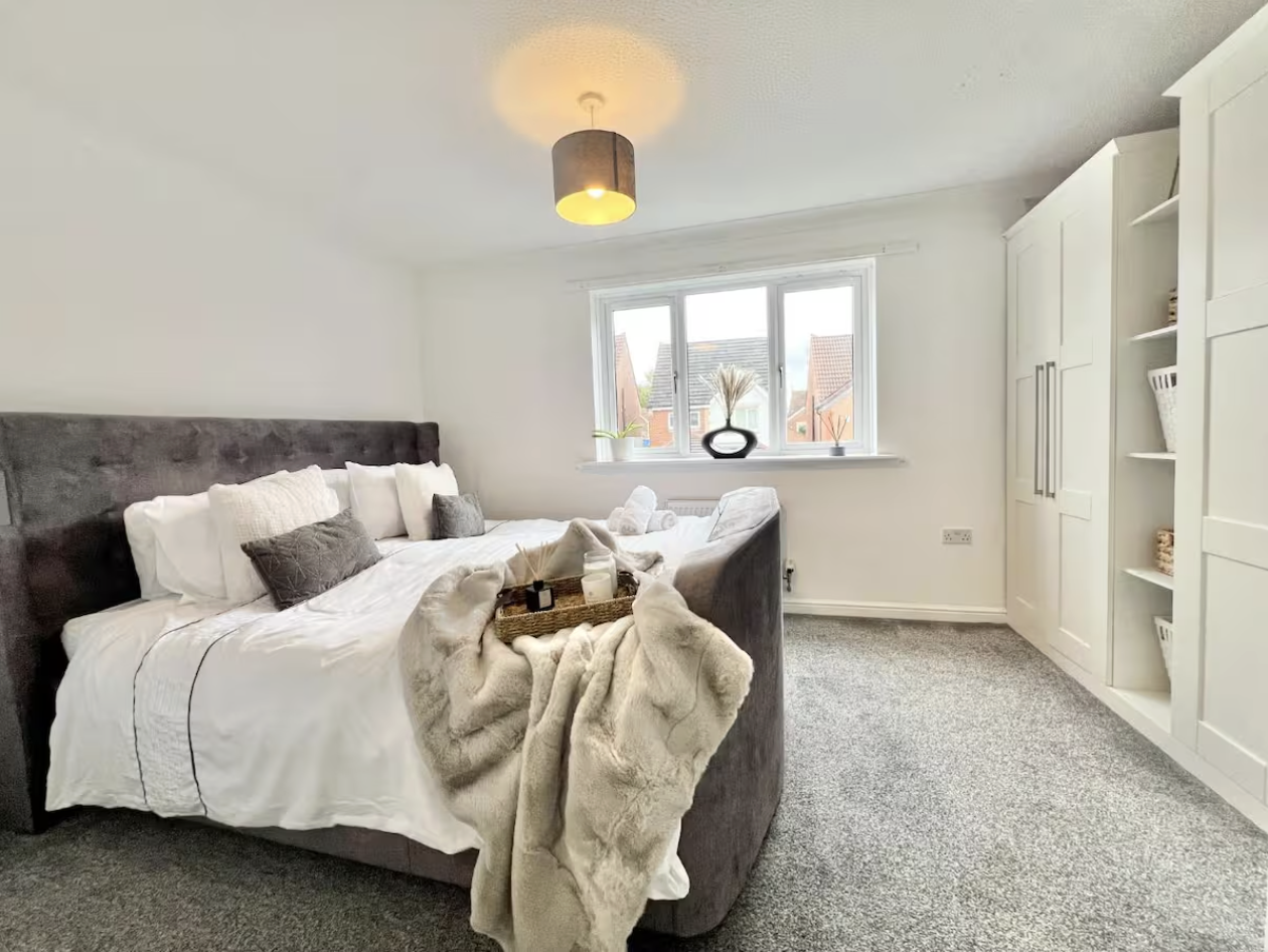 A cozy bedroom with a large bed, white bedding, and gray pillows. A tray with candles and decor is on a beige blanket at the foot of the bed. A window with a sill holds a black vase with dried flowers and a plant. White built-in shelves and a closet are on the right, and a ceiling light hangs above.