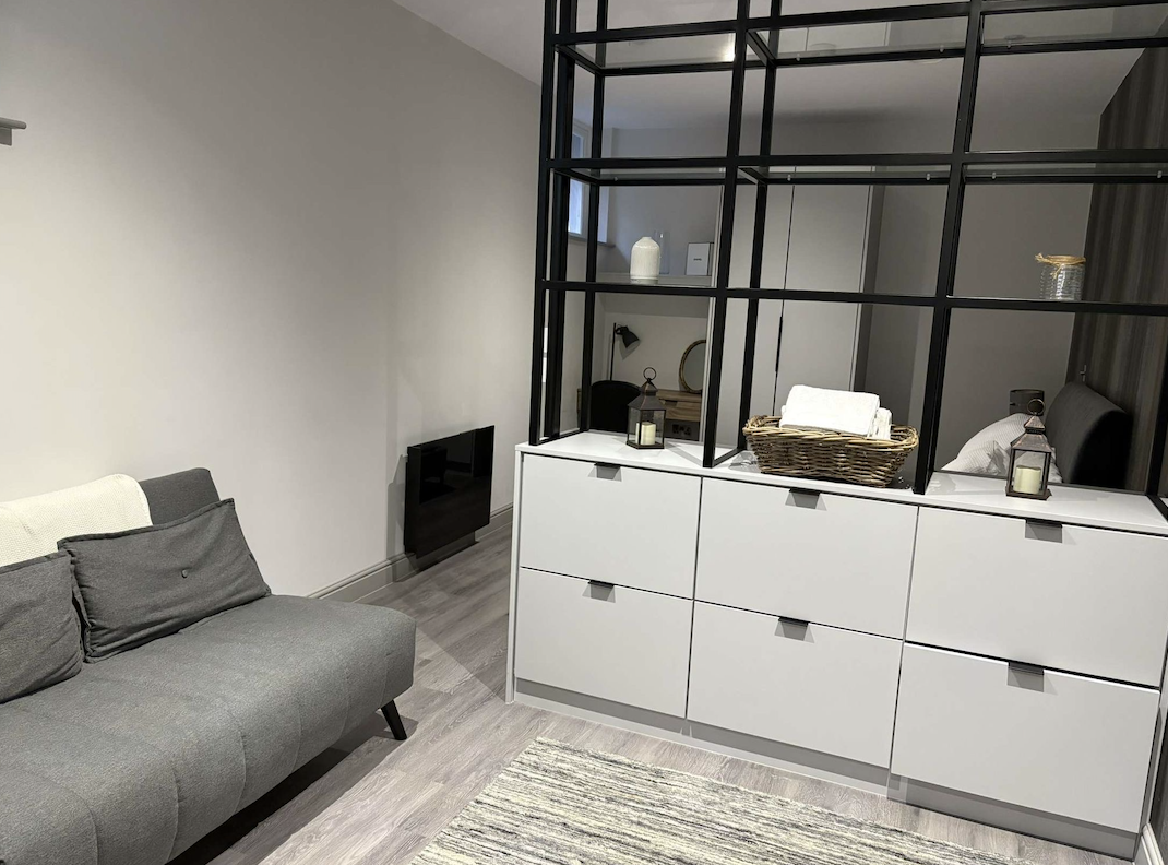 Living room with gray sofa, black room divider, white dresser with baskets and candles, and a small wall-mounted heater.