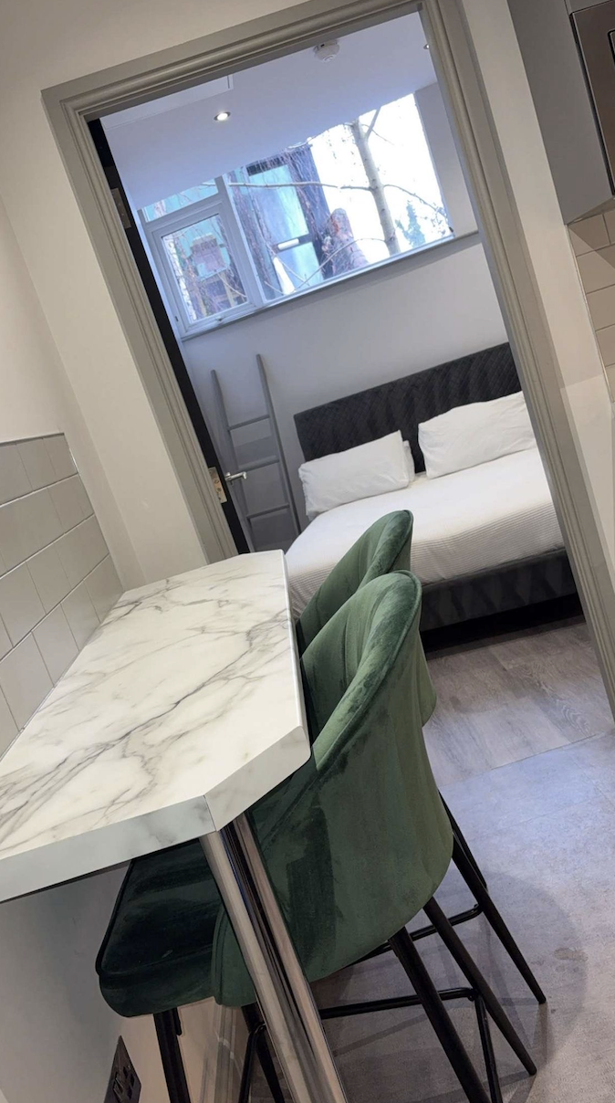 A view through a doorway showing a small living area with a marble table, green velvet chairs, a bed with white pillows, a window, and a person taking a photo outside.