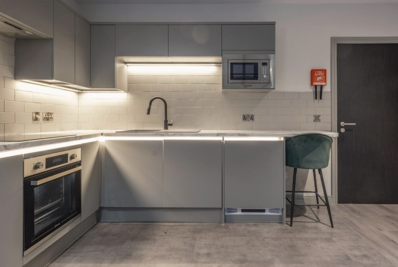 Modern kitchen with gray cabinets, white subway tile backsplash, built-in oven, microwave, black faucet, countertop with under-cabinet lighting, a green bar stool, and a dark door with a fire safety device on the wall.