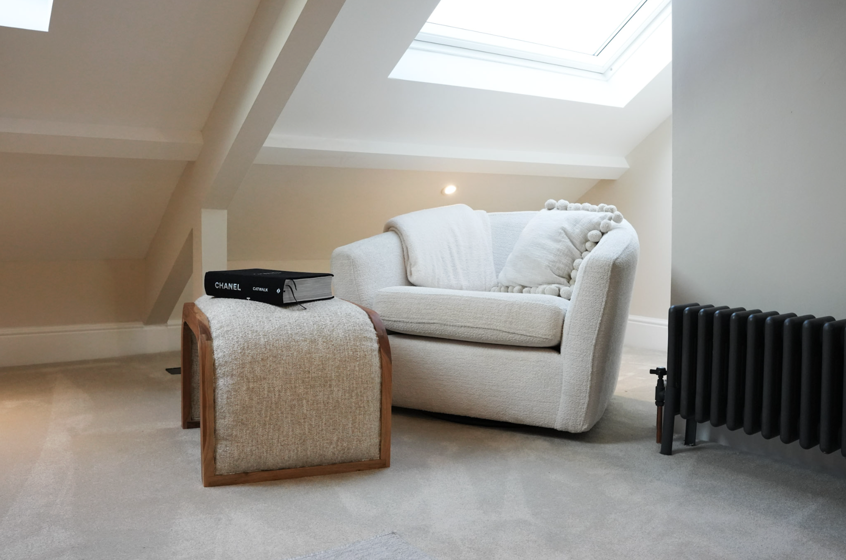 A white armchair with a matching pillow and throw blanket, a small wooden and fabric ottoman with a black book titled 'Chanel' on top, a black radiator, and a skylight window in a neutral-colored attic space.