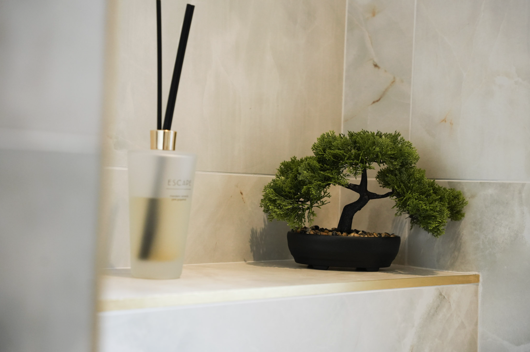 A small bonsai tree in a black pot and a white diffuser with black reeds on a beige tiled shelf.