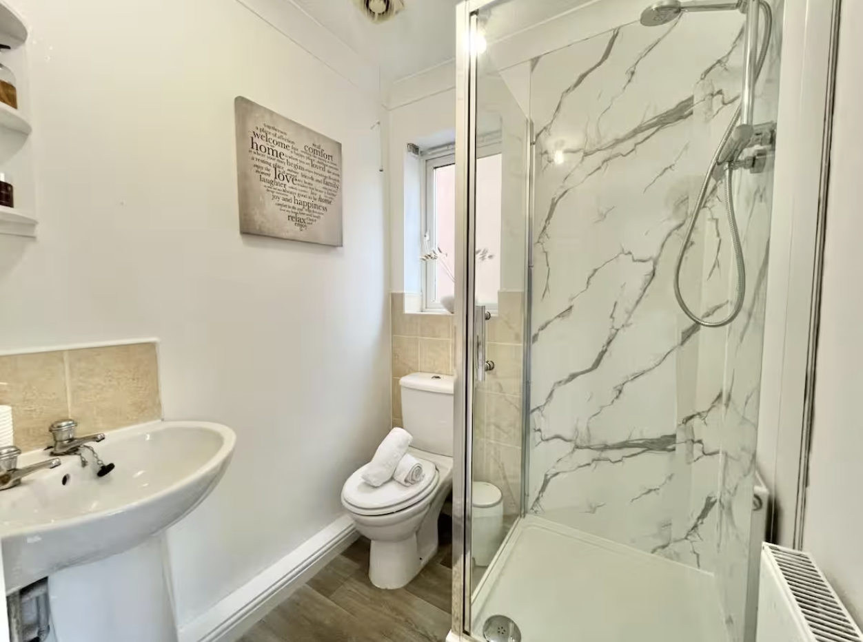 Small bathroom with a pedestal sink, a toilet with towels on it, a shower with marble-like walls, a small window, and a wall decoration.