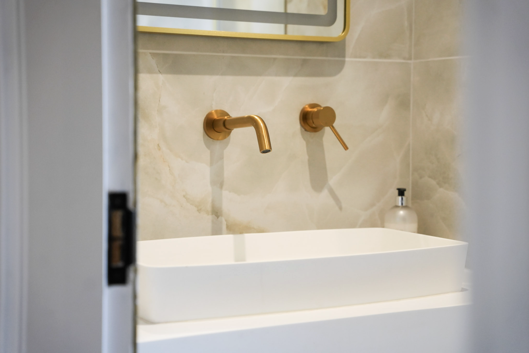 Bathroom sink with a gold faucet and a soap dispenser in the background, viewed through a partially opened door.