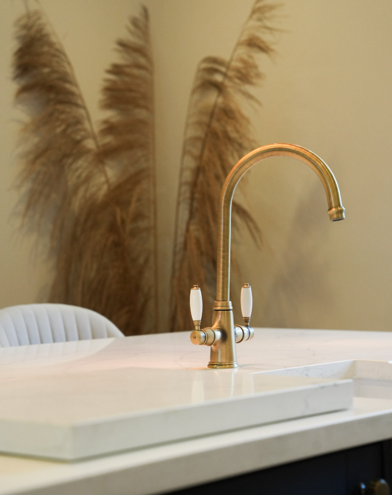 Close-up of a modern kitchen sink with a brushed gold faucet on a white marble countertop, with tall beige pampas grass in the background.