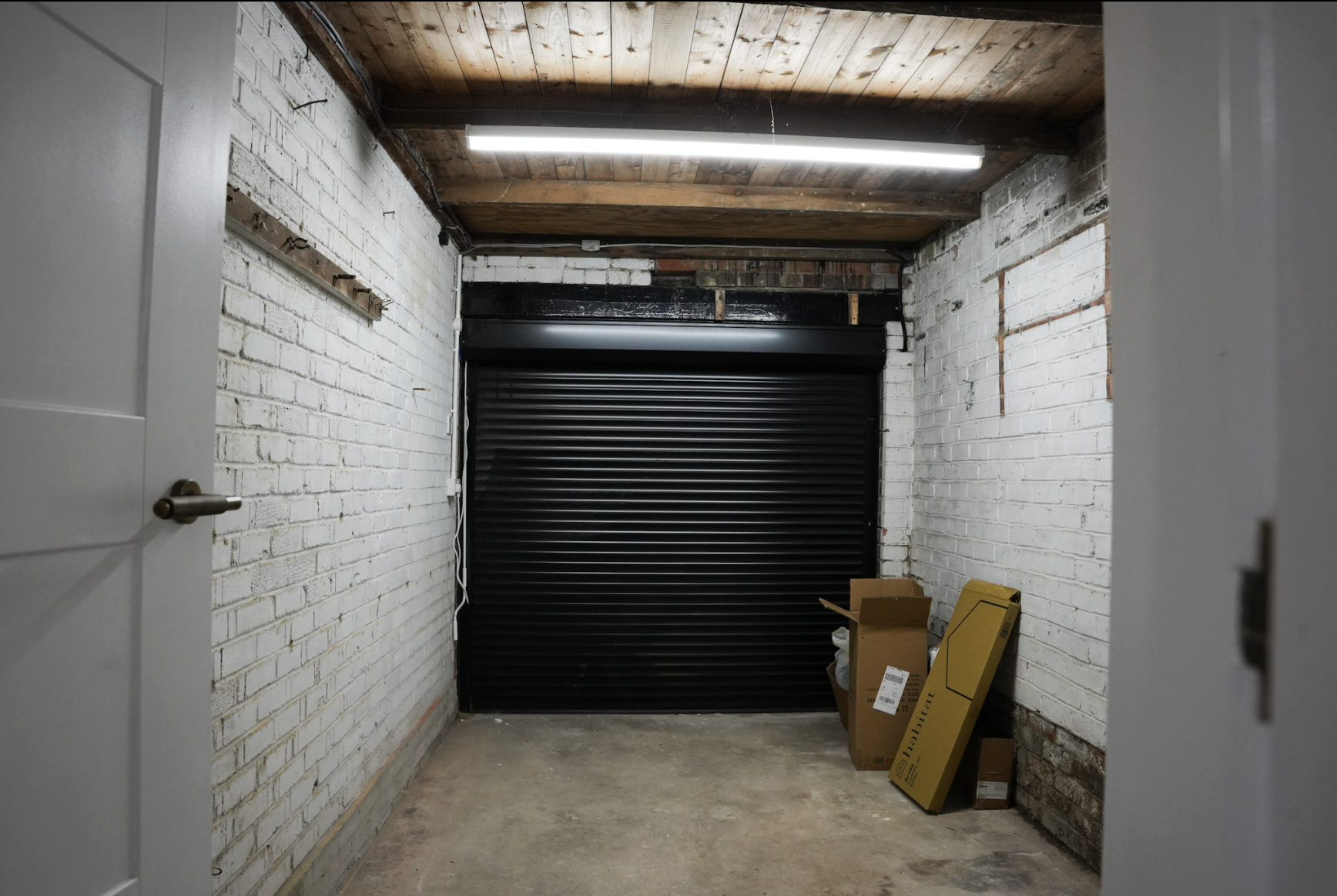 Underground storage unit with white brick walls, a concrete floor, and a wooden ceiling with fluorescent lighting. There is a black roll-up garage door at the back and a few cardboard boxes on the right side.