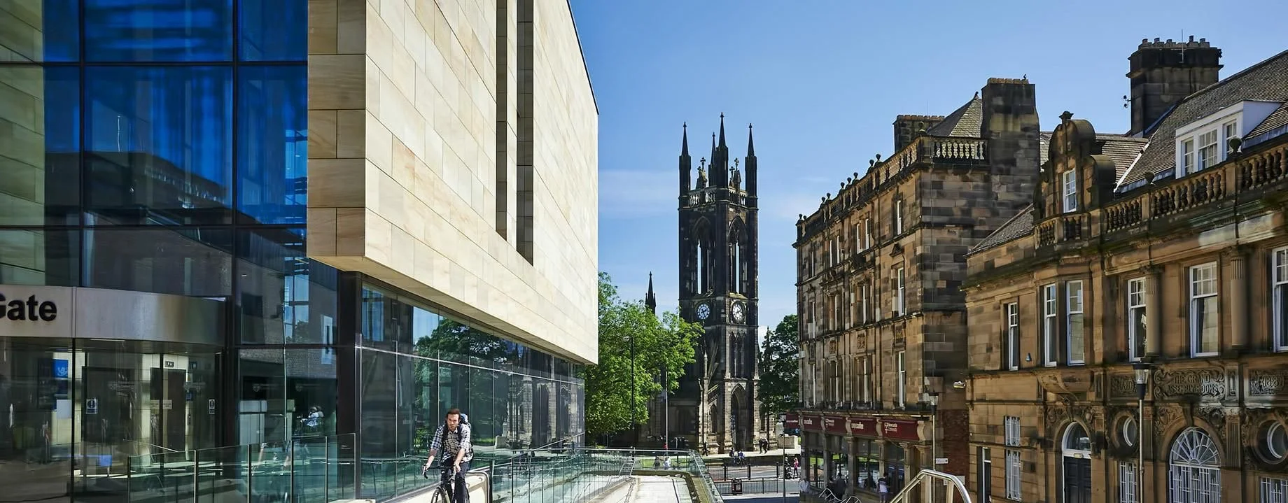 A city street scene with a modern glass building on the left and older historic buildings on the right, featuring a tall church steeple in the background, and a man riding a bicycle on the sidewalk.