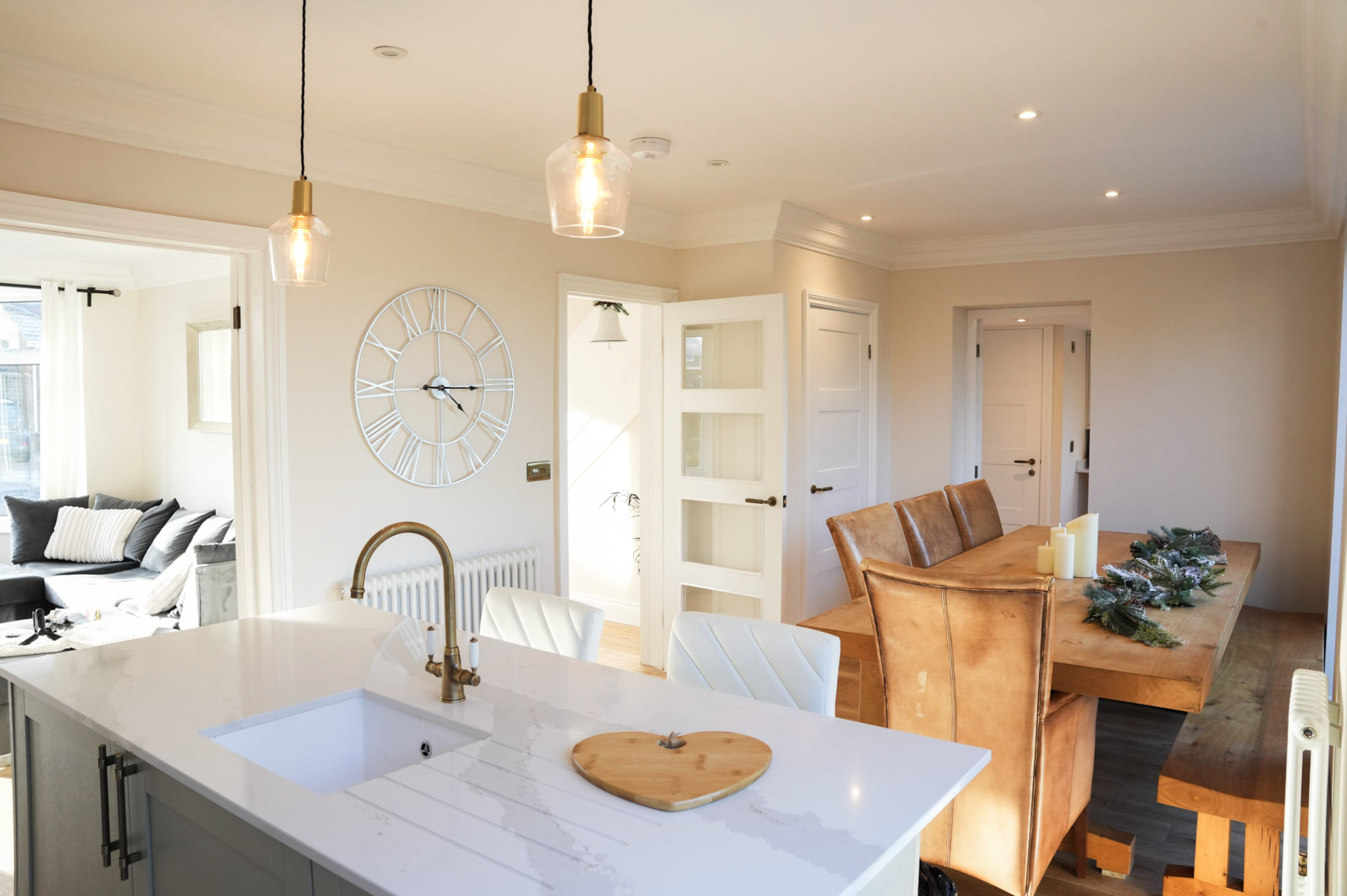 Modern open-concept kitchen and dining area with white countertops, brown leather chairs, a wooden dining table with candles and pine decoration, and a large wall clock.