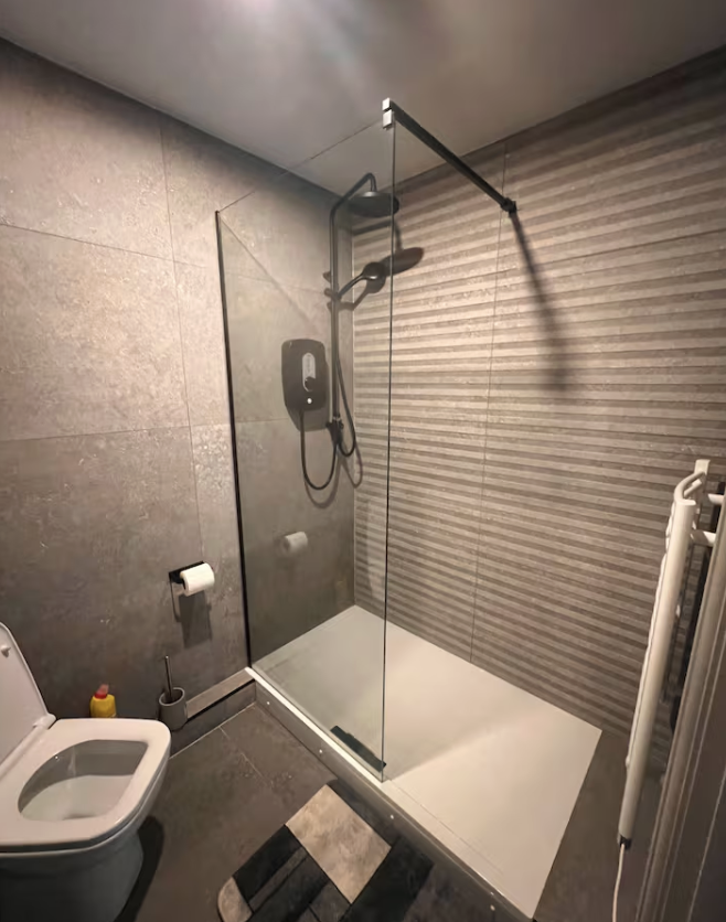 A modern bathroom with a walk-in shower featuring a glass door, a shower head, and a black shower unit on the wall. To the left is a toilet, and on the right a white towel radiator. The floor and walls are tiled in gray, with a small rug in front of the shower.