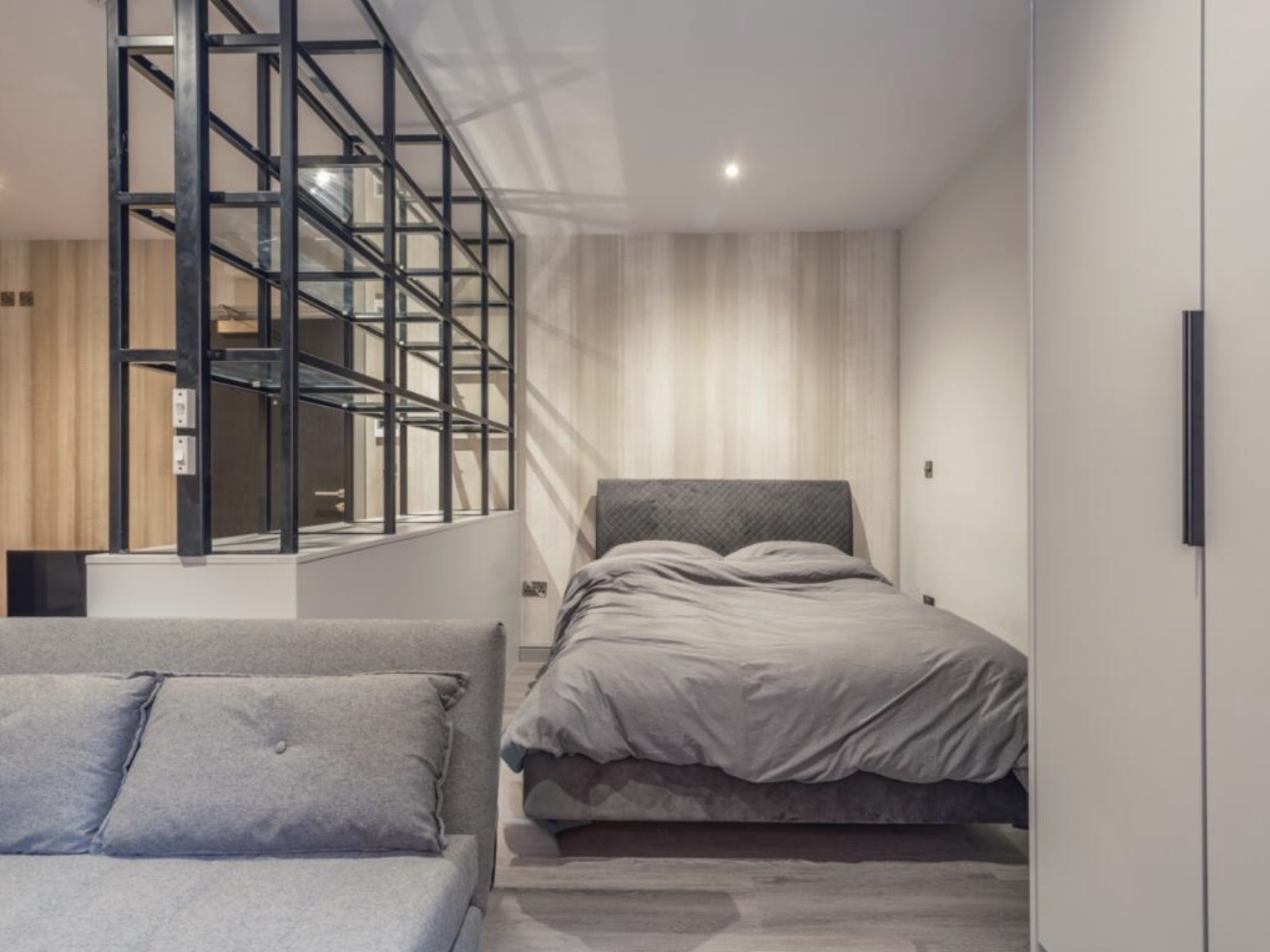 Modern bedroom with gray bed and headboard, gray sofa, and white walls, separated by a black metal grid partition.
