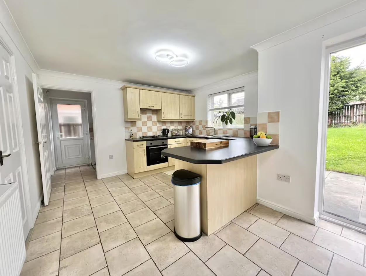 A bright kitchen with beige cabinets, a black countertop, a gas stove, and a window with a potted plant. There is a door leading outside to a grassy backyard.