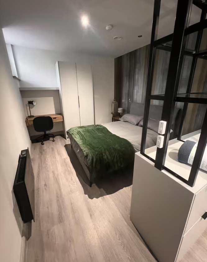 A modern bedroom with a bed covered in a green blanket, positioned against a wooden accent wall. To the left of the bed, a small nightstand with a lamp. On the far left, a desk with a black swivel chair and a desk lamp. There is a white wardrobe against the wall near the ceiling. The room has light-colored wood flooring and a black wall heater along the left wall.