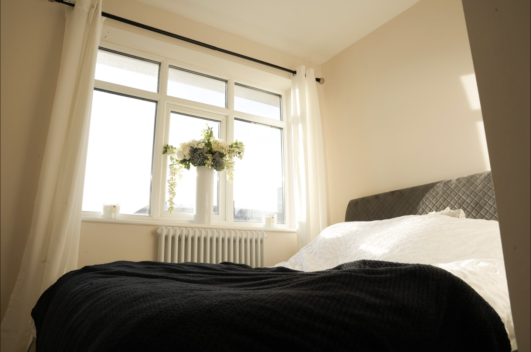 A bedroom with a large window, cream-colored walls, a bed with a white cover and gray headboard, a black blanket at the foot of the bed, and a tall white vase with a bouquet of white and gray flowers on the windowsill. Light curtains frame the window, and there are small candles or decorative objects on the window sill.