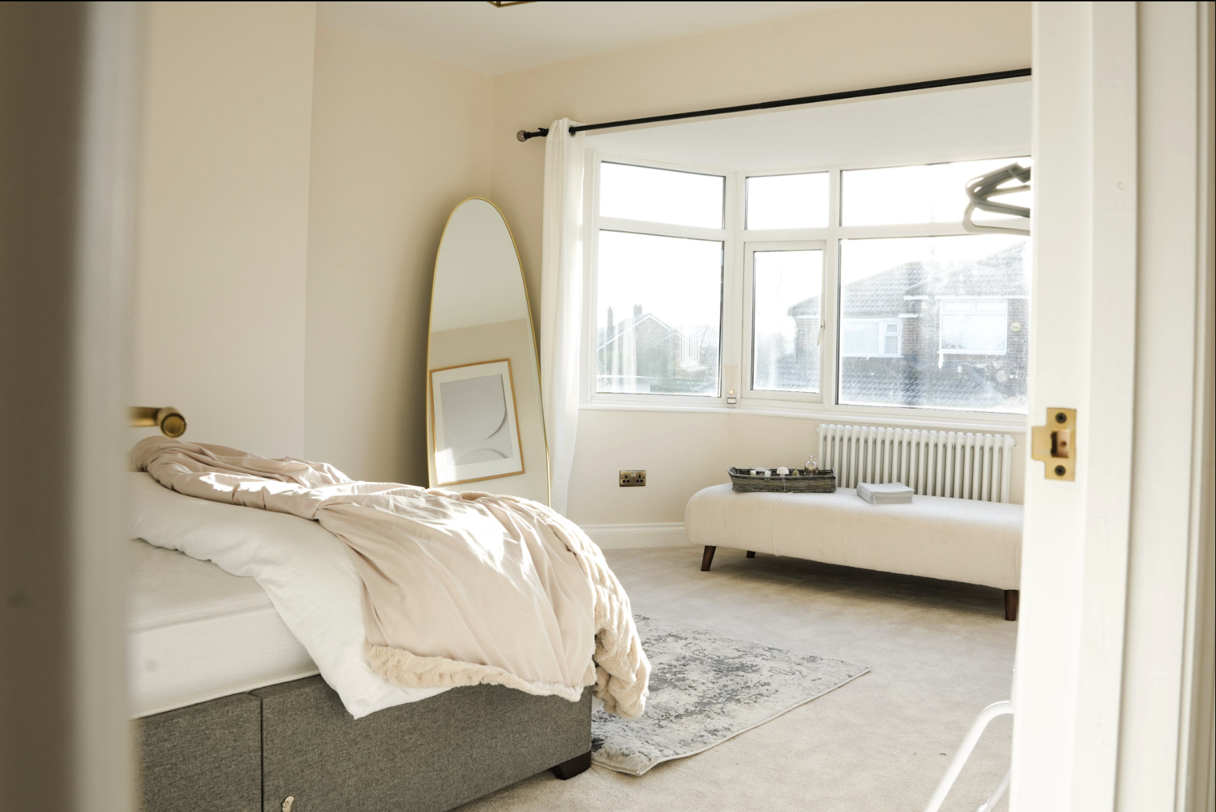 A bright bedroom with a bed on the left, a large mirror leaning against the corner, a window with daylight coming in, and a bench underneath the window with a tray and books on top.