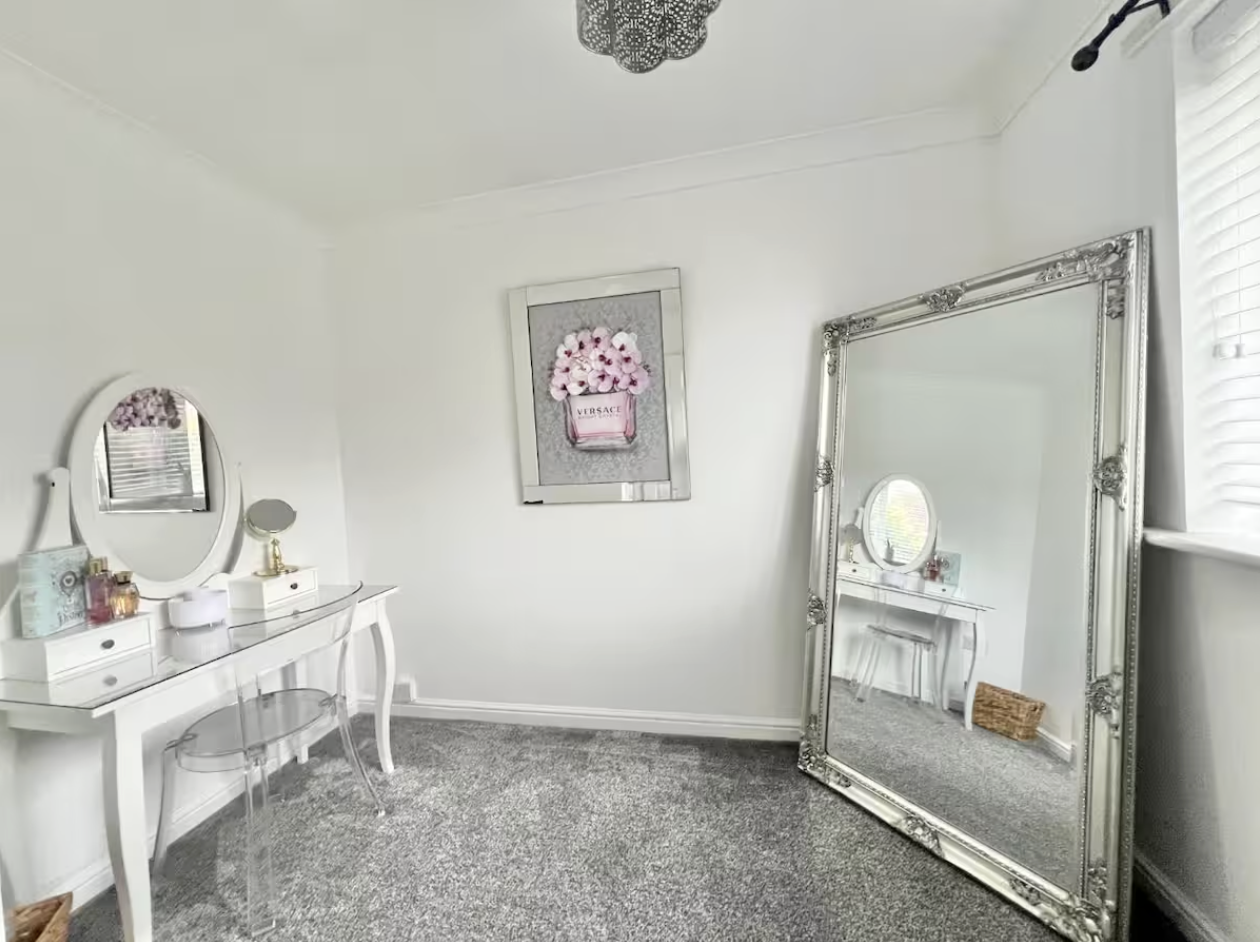 A white bedroom with a large ornate mirror, a small vanity table with a round mirror, and a framed picture of pink flowers and Versace branding on the wall; the room has gray carpet and a window with blinds.