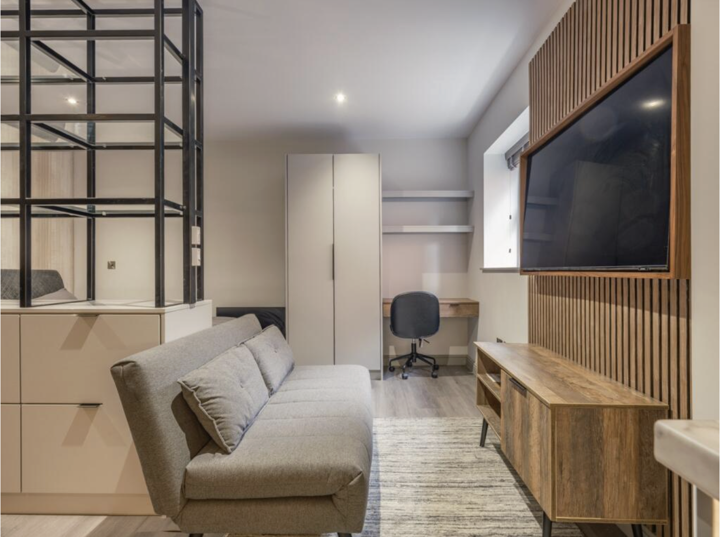 Modern living room with a beige sofa, wooden media console, black wall-mounted TV, small workspace with a chair and desk, and a white wardrobe next to a window.