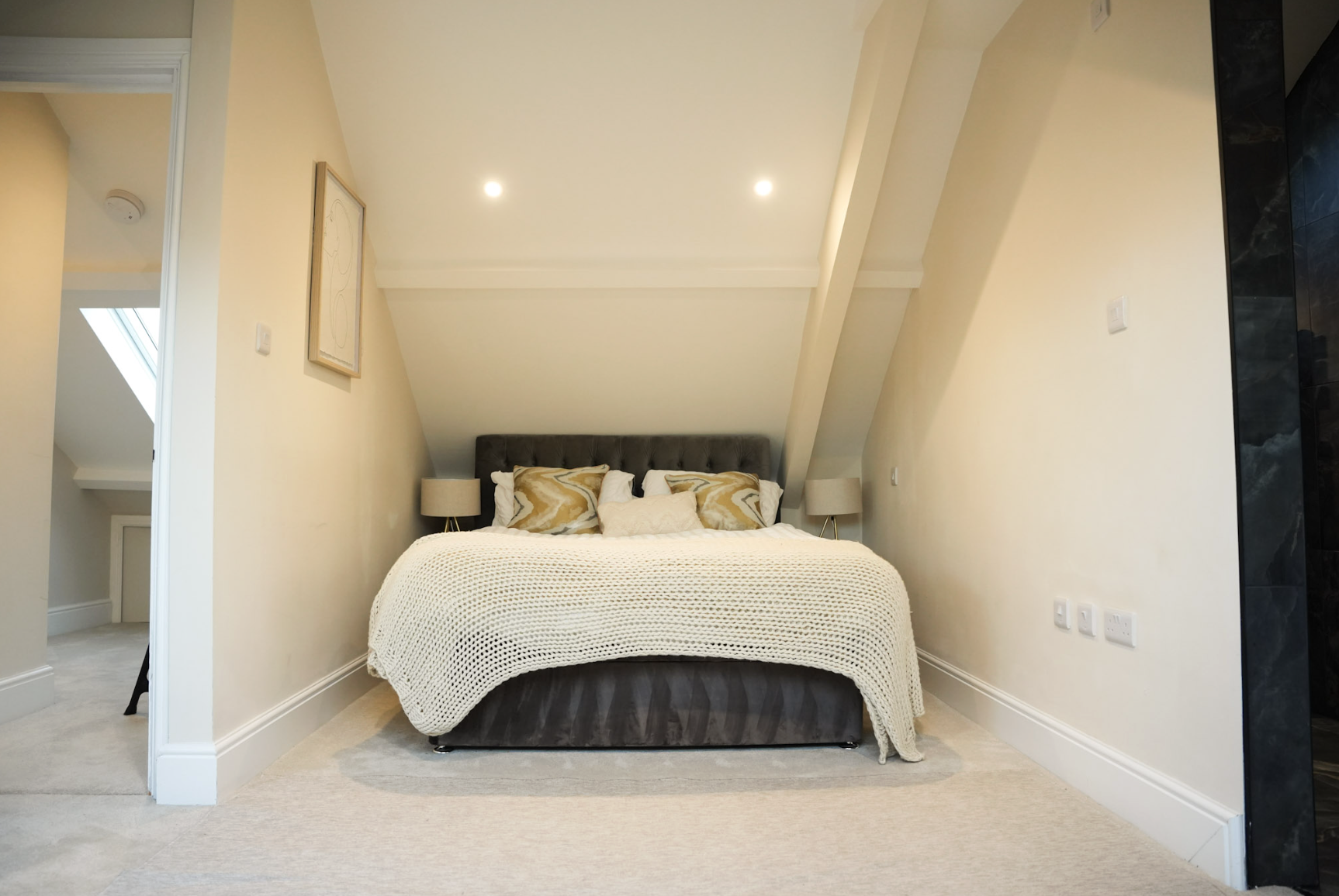 A bedroom with a bed, pillows, and lamps, located under a sloped ceiling with recessed lighting.