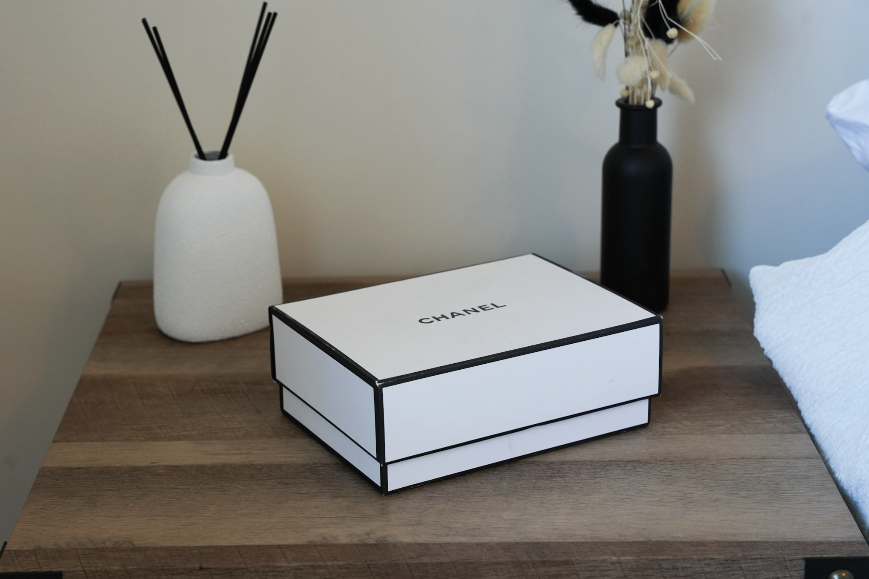 A black and white Chanel shoebox on a wooden bedside table, with white and black vases containing decorative dried flowers and reeds nearby.