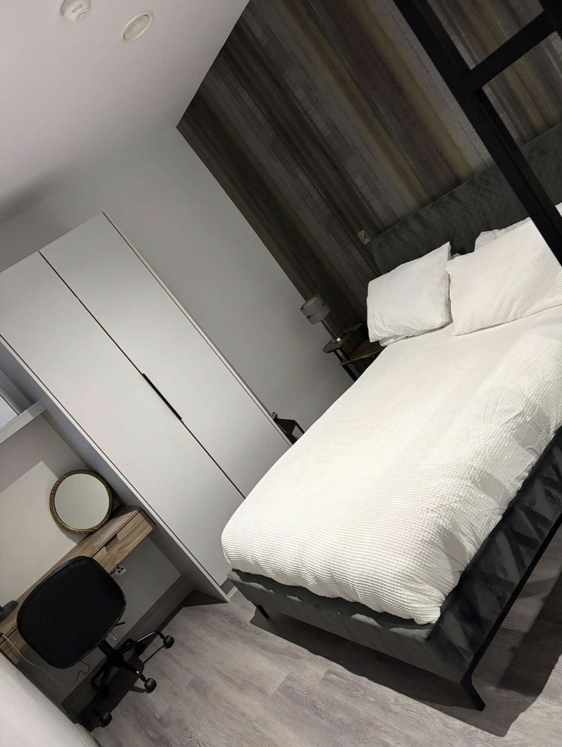 A modern bedroom with a bed, white bedding, and two pillows. There's a black metal headboard, a dark wood accent wall behind the bed, and a white wardrobe. A small side table with a lamp and a black office chair at a study desk with a mirror and a wooden drawer are also visible.