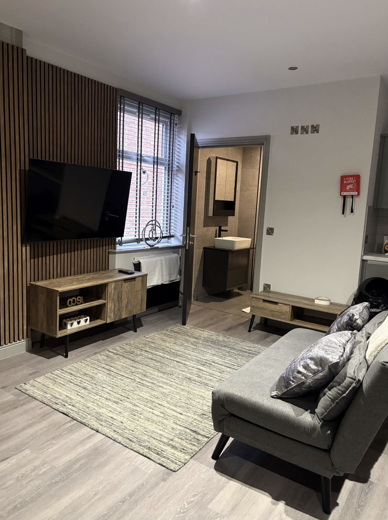 Modern living room with a grey sofa, metallic pillows, wooden media console, wall-mounted flat-screen TV, window with blinds, a small rug, and an open door leading to a washroom area.