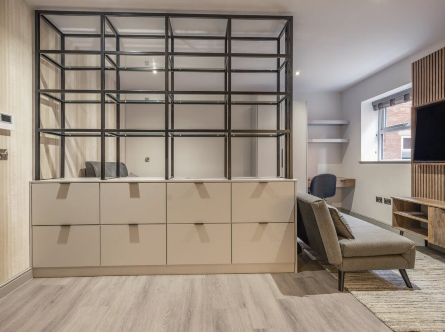 Living room with beige storage unit, glass cube shelf, gray armchair, black office chair at wooden desk, and hardwood floor.