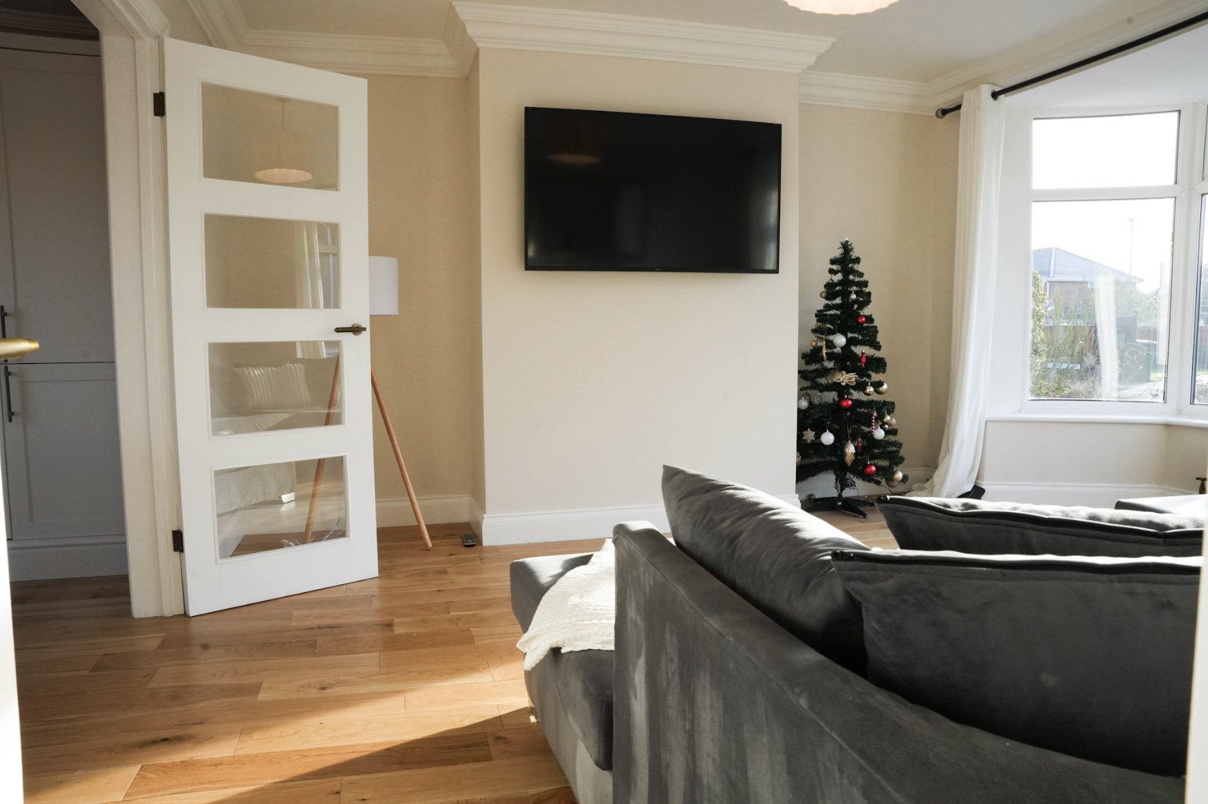 Living room with a sectional sofa, a wall-mounted flat-screen TV, a small decorated Christmas tree near a large window with white curtains, and a partially open white door with glass panes.