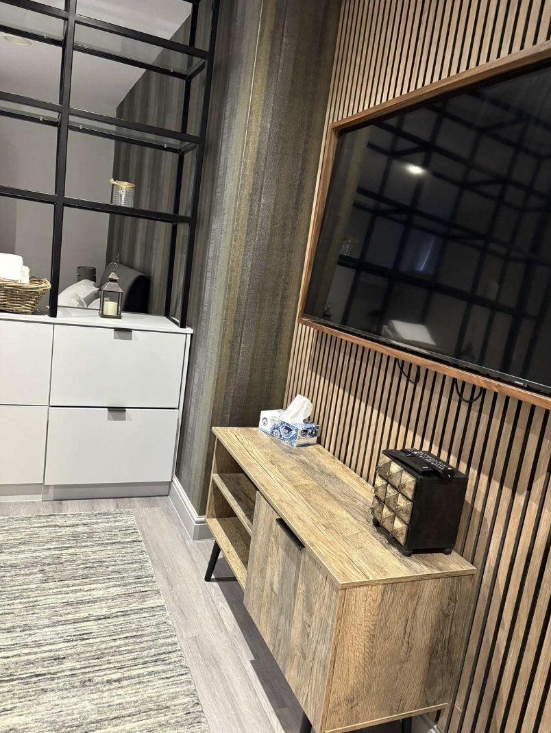 A modern living room corner with a large flat-screen TV on a wood-paneled wall, a wooden media console, and a white cabinet with shelves. Decor includes a small lantern, a basket, and a tissue box.