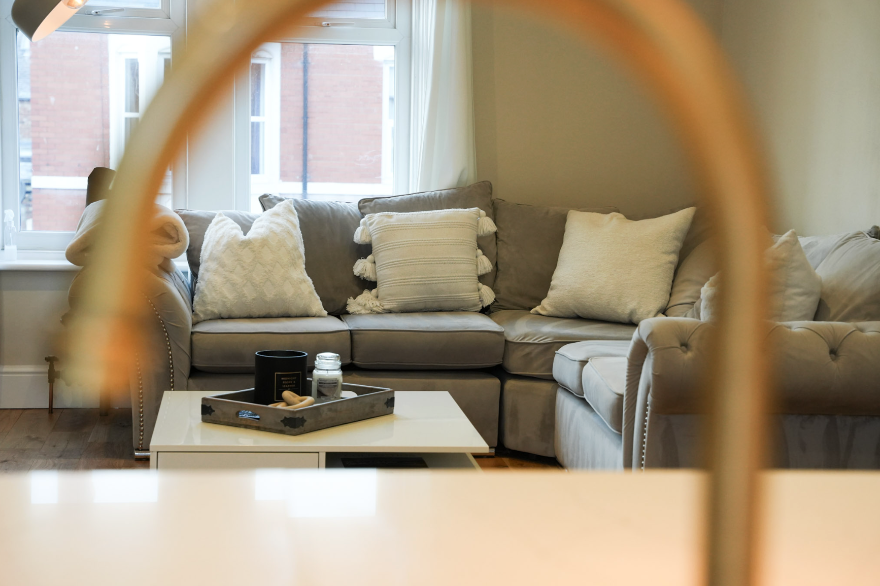 Living room with a beige sectional sofa decorated with white and cream throw pillows, a white coffee table with decorative items, large windows with curtains, and a view of a red brick building outside.