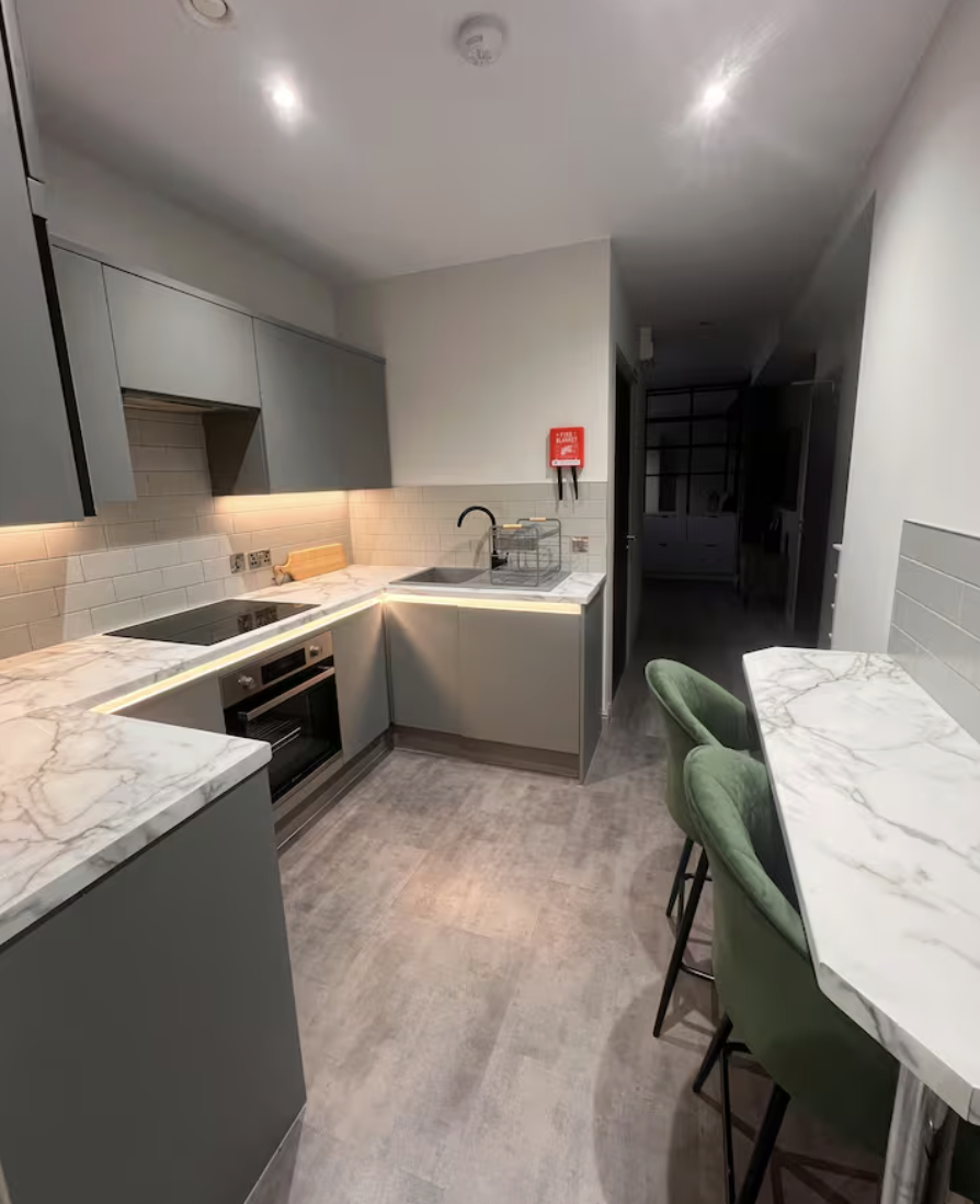 Modern kitchen with gray cabinets, marble countertops, and a tiled backsplash, featuring a stove, oven, sink, and green barstool chairs.