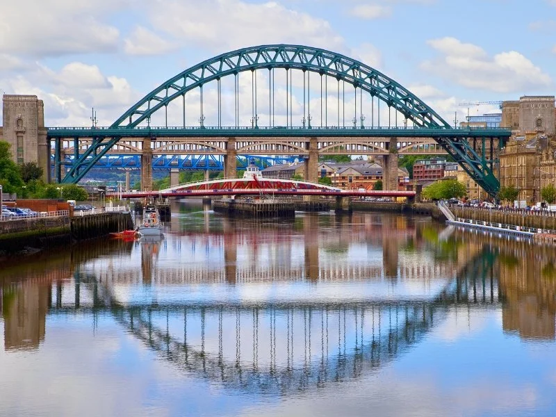 A cityscape featuring two bridges crossing a river, with buildings and a partly cloudy sky in the background.