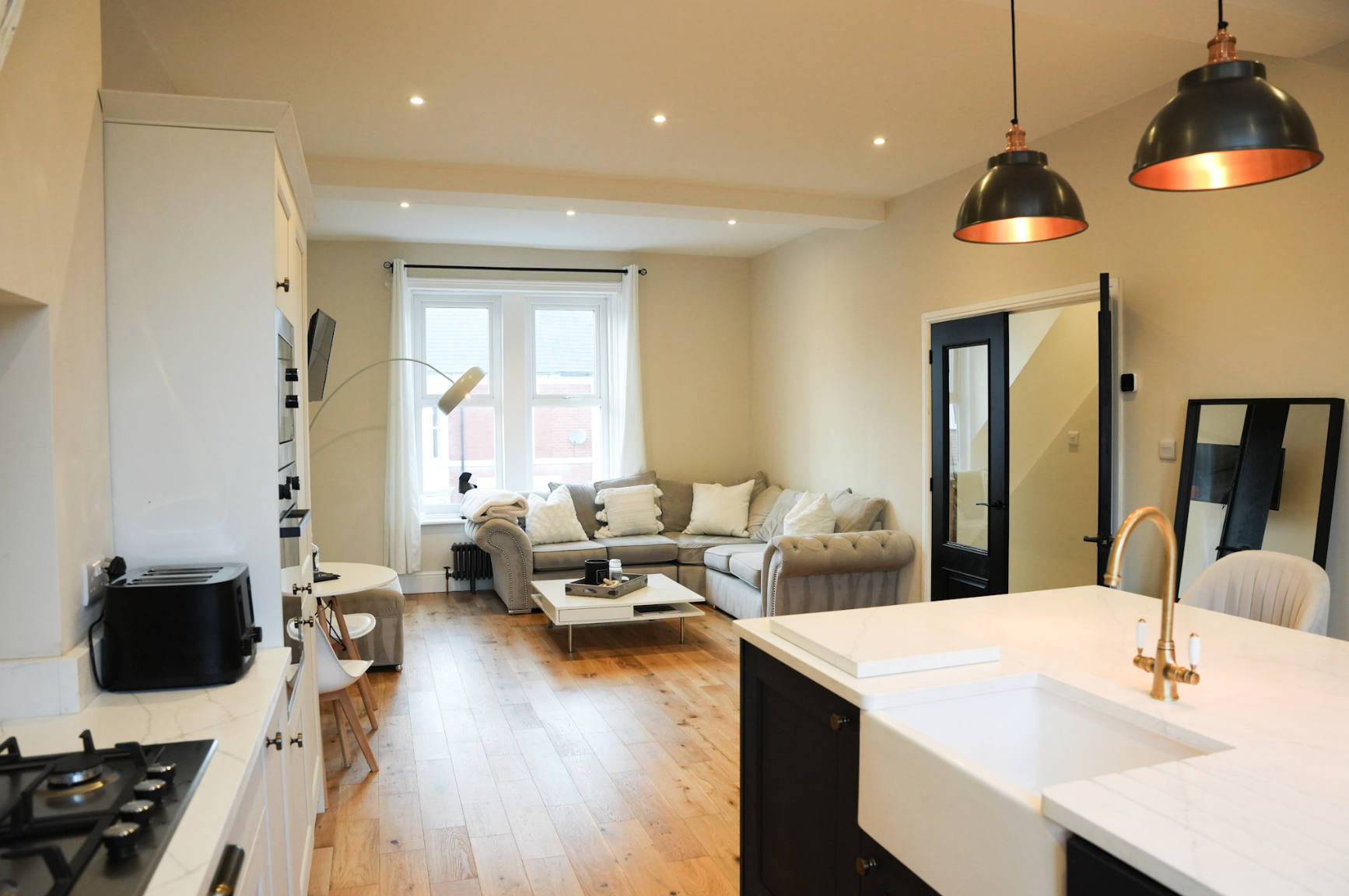 Open-concept living room and kitchen with beige sectional sofa, white coffee table, hardwood floors, black door, black and copper pendant lights, white kitchen island with brass faucet, and large window with white curtains.