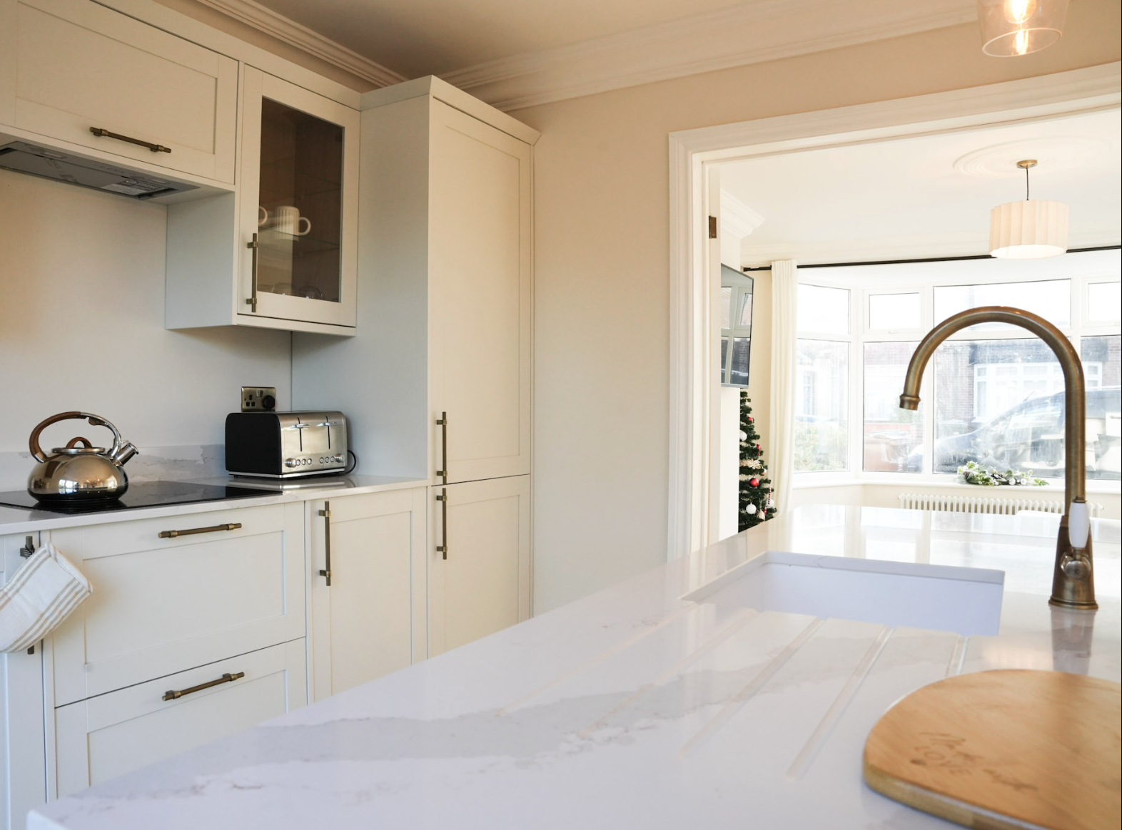 Bright kitchen with white cabinets, a tiled countertop, a stainless steel kettle and toaster on the counter, and a large window area with a Christmas tree inside, indicating a holiday season.