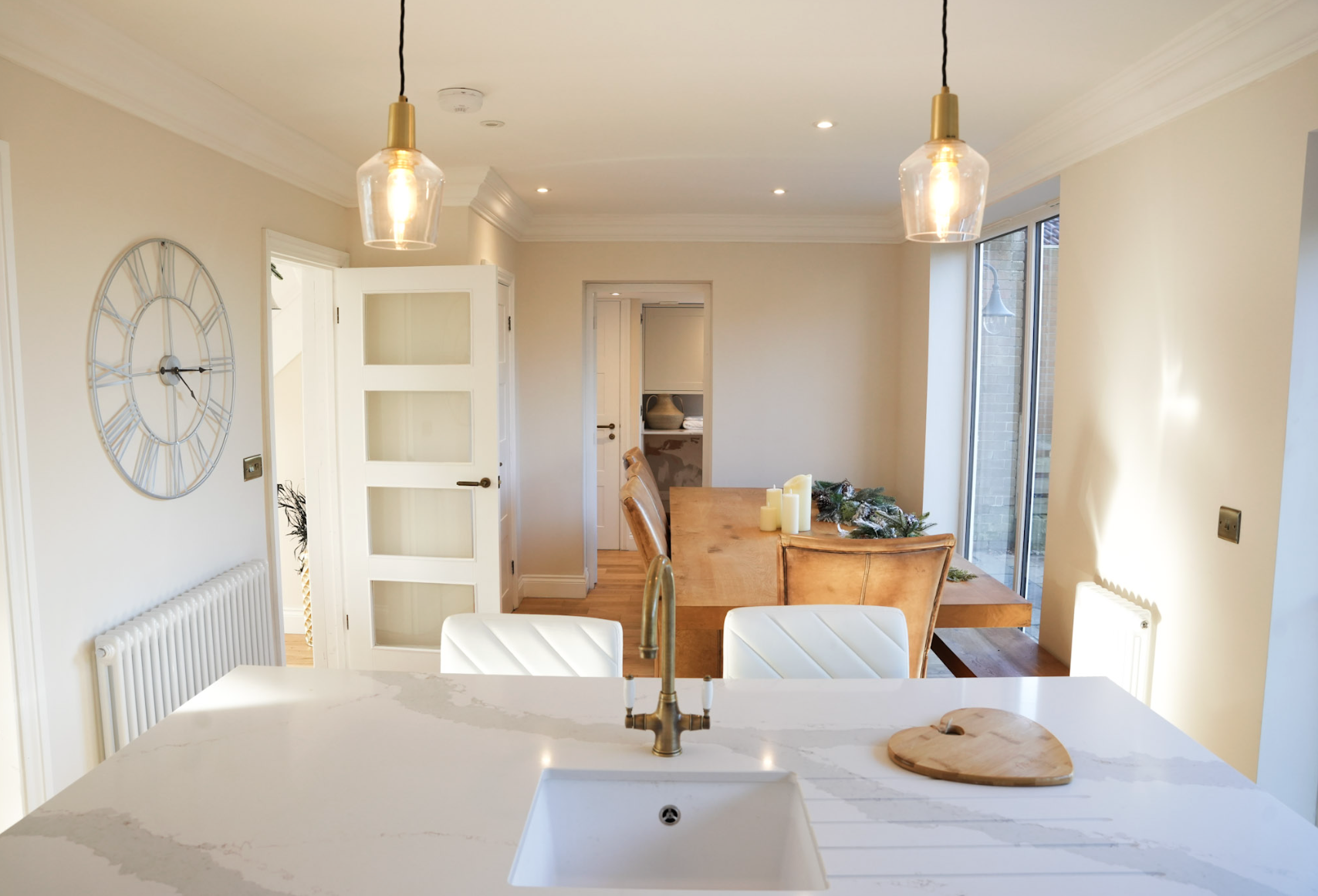 Bright, modern kitchen and dining area with a white marble island, gold pendant lights, a wooden dining table decorated with candles and greenery, a large clock on the wall, and sliding glass doors leading outside.