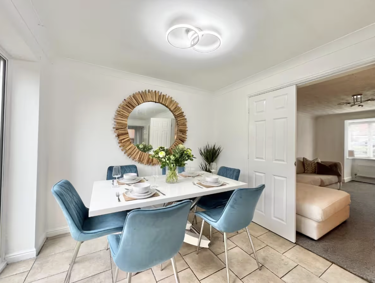 A dining room with a white table, six blue velvet chairs, a vase with flowers, and a round wooden-framed mirror on the wall. Part of an adjacent living room with a beige sectional and gray carpet is visible.