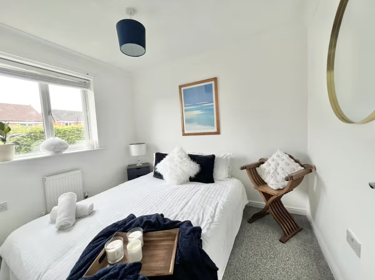 A neatly made bed with white bedding, decorative pillows, and bath towels on top. A wooden tray with candles and a blanket is on the bed. There is a window with a potted plant and a decorative object on the sill, a small nightstand with a lamp, a framed art piece of a beach scene, a hanging blue ceiling light, and a wooden chair with a patterned cushion in a bright, cozy bedroom.