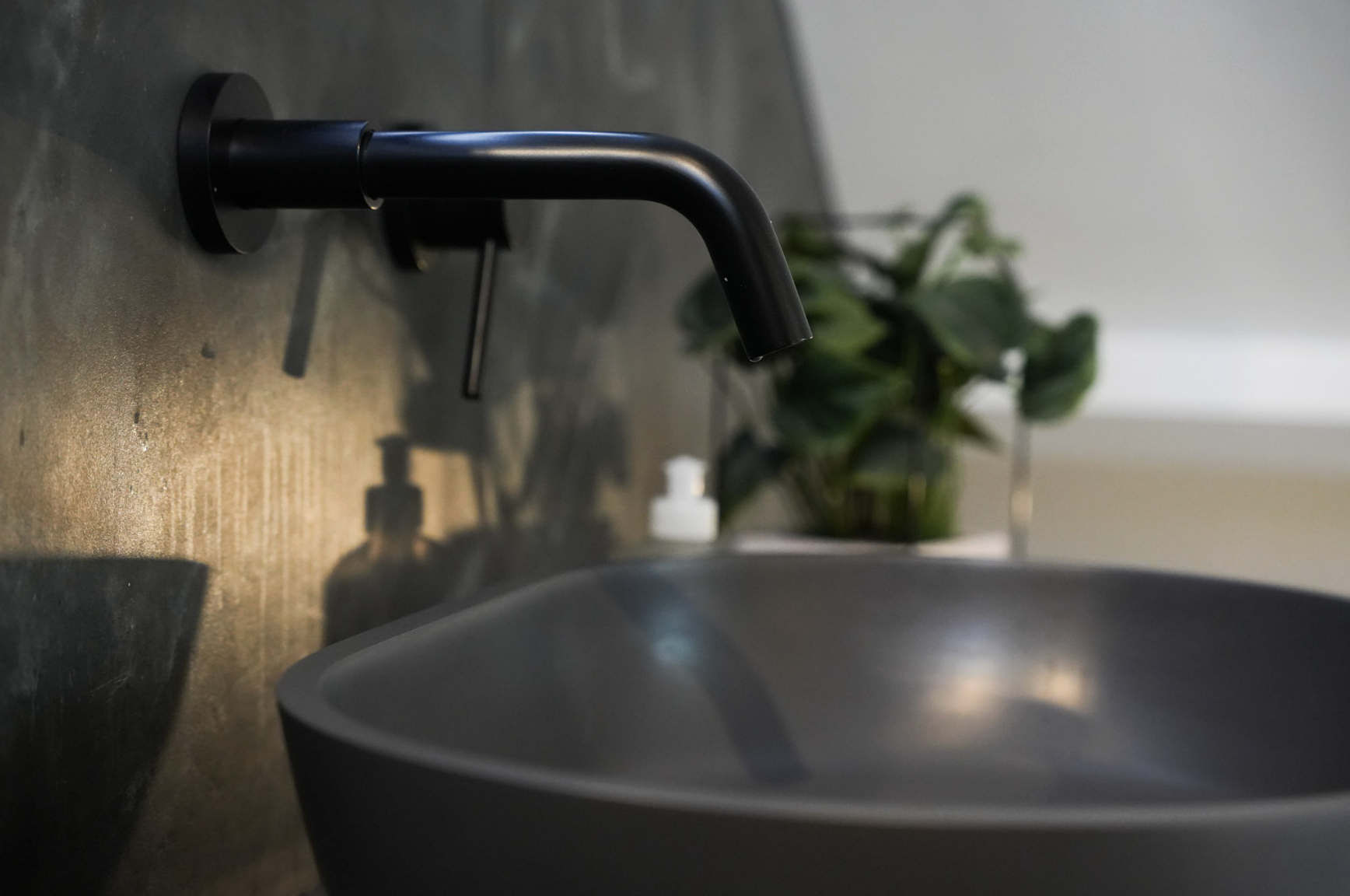 Black bathroom faucet above a gray sink with a blurring green plant in the background.