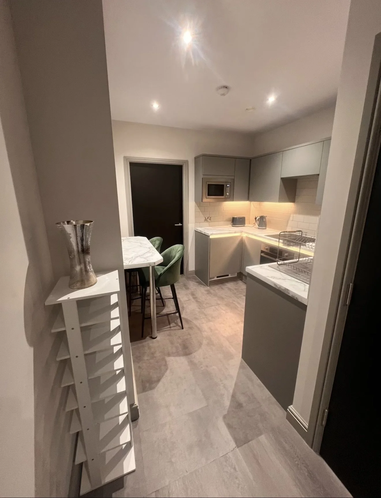 A modern kitchen with gray cabinets, white marble counters, and a small dining area with green chairs. Recessed lighting illuminates the space, which features a microwave, toaster, kettle, and dish rack.