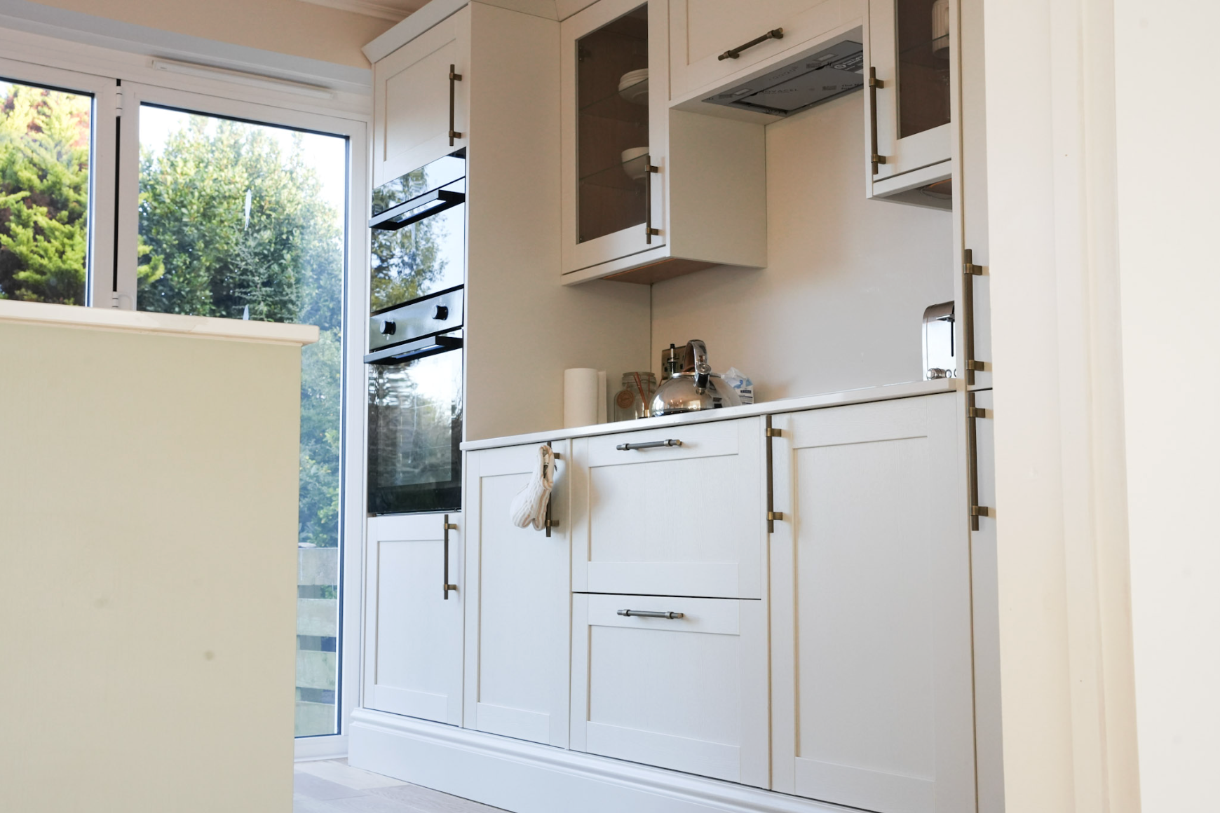 White kitchen cabinets with gold handles, a stainless steel kettle, paper towel roll, and a window showing trees outside.