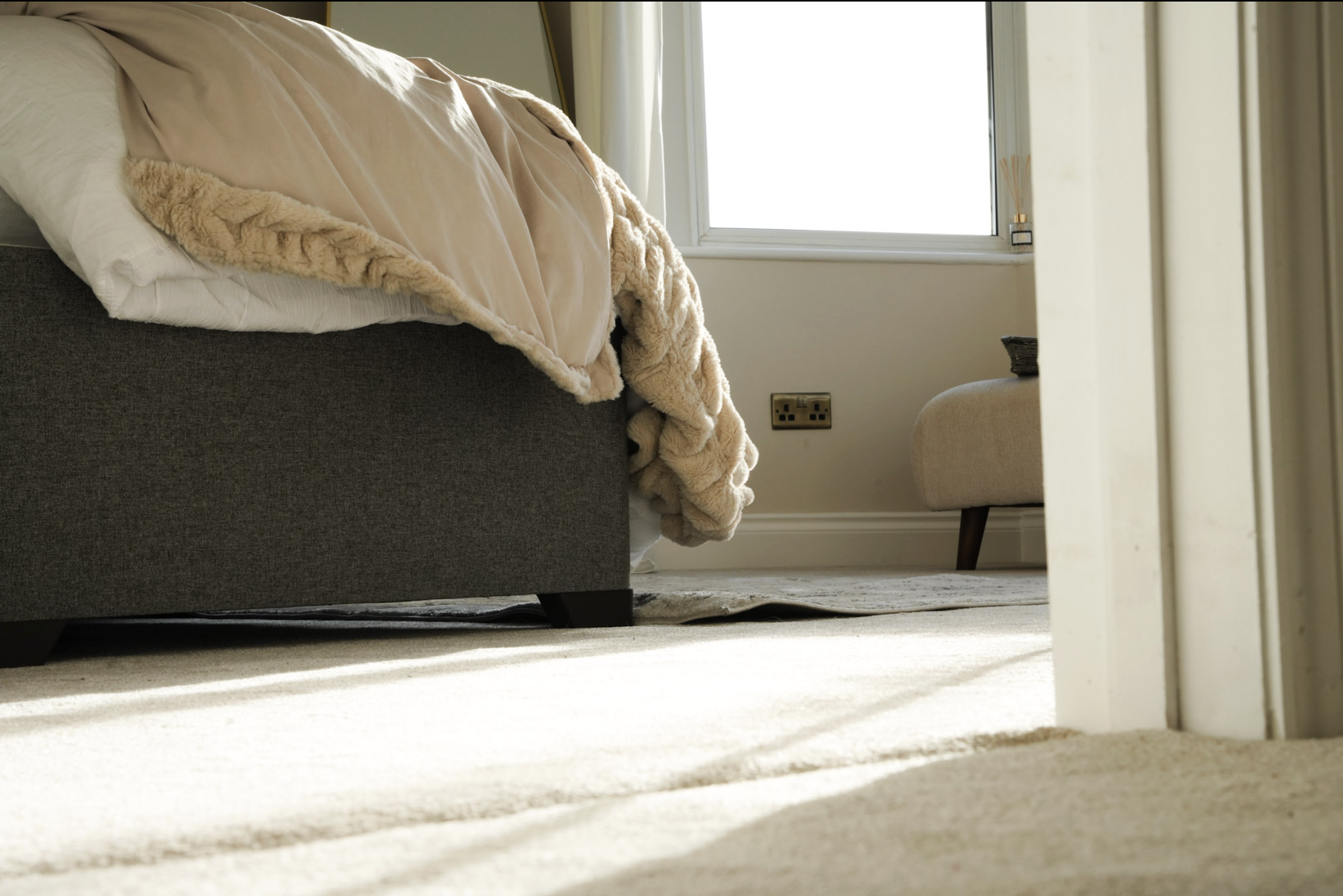 A bed with beige and cream-colored bedding, partially made, in a bedroom near a window with sunlight.