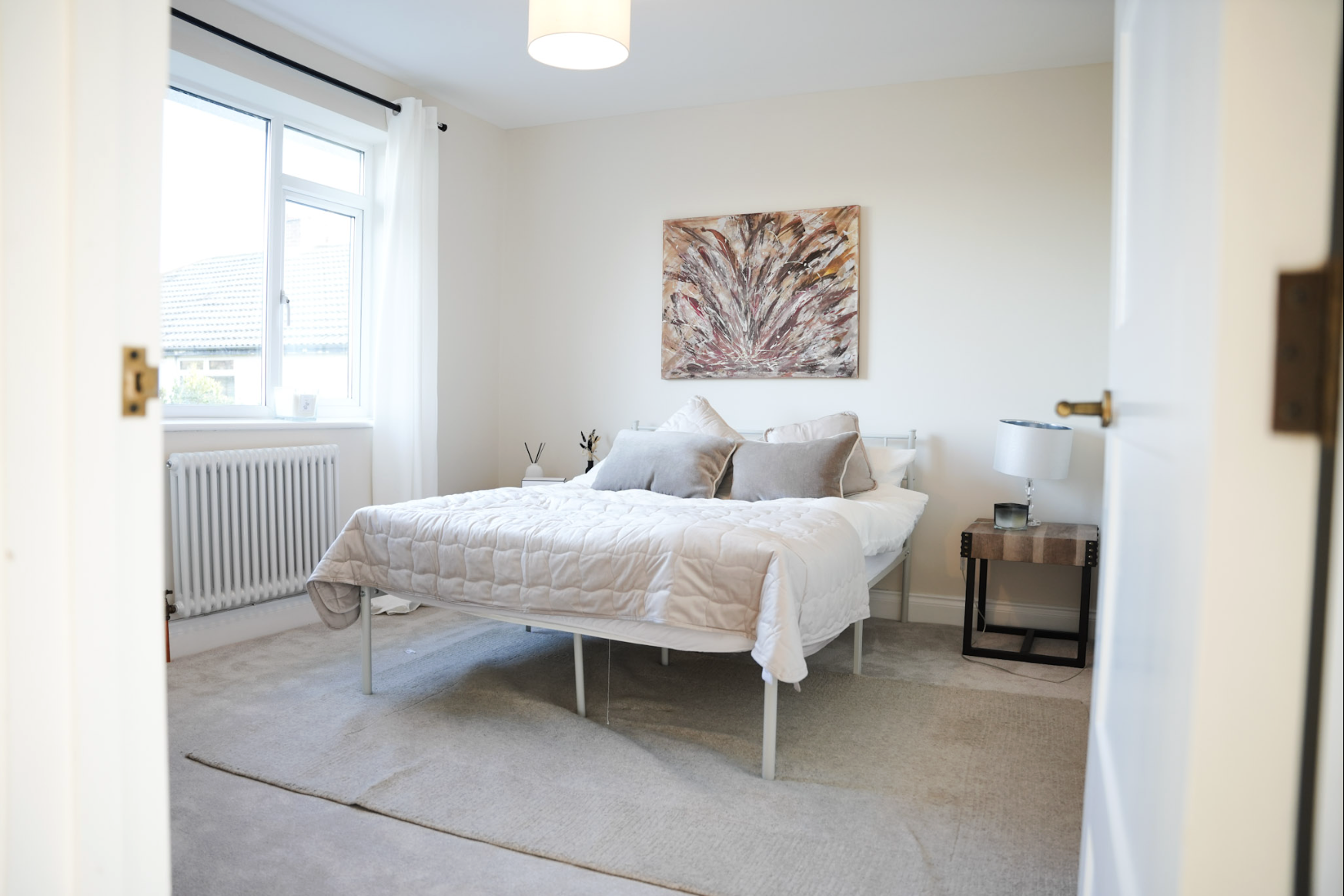 Photo of a bedroom seen through an open door, featuring a bed with white bedding and several pillows, a window with white curtains, a side table with a lamp and picture frame, and a large abstract painting on the wall.