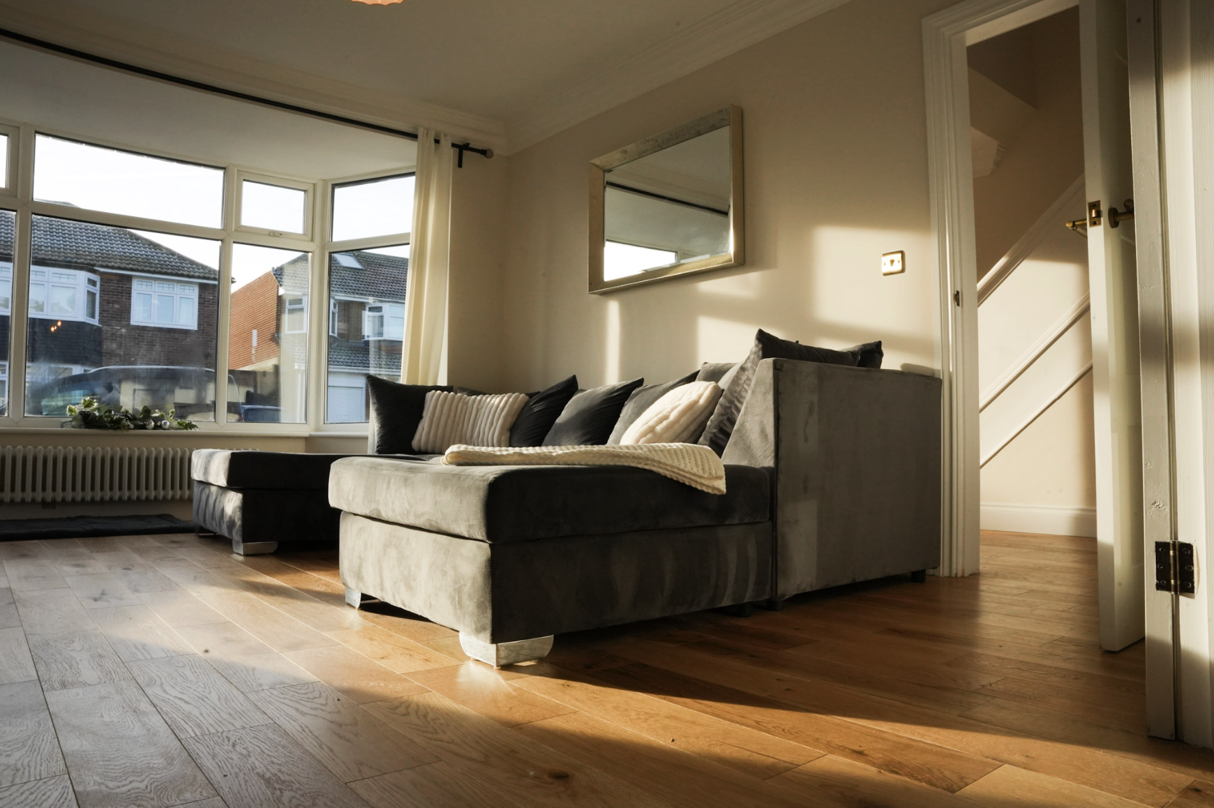 Sunlit living room with a gray sectional sofa, black and white throw pillows, a wall mirror, large windows with cream curtains, and wooden flooring.