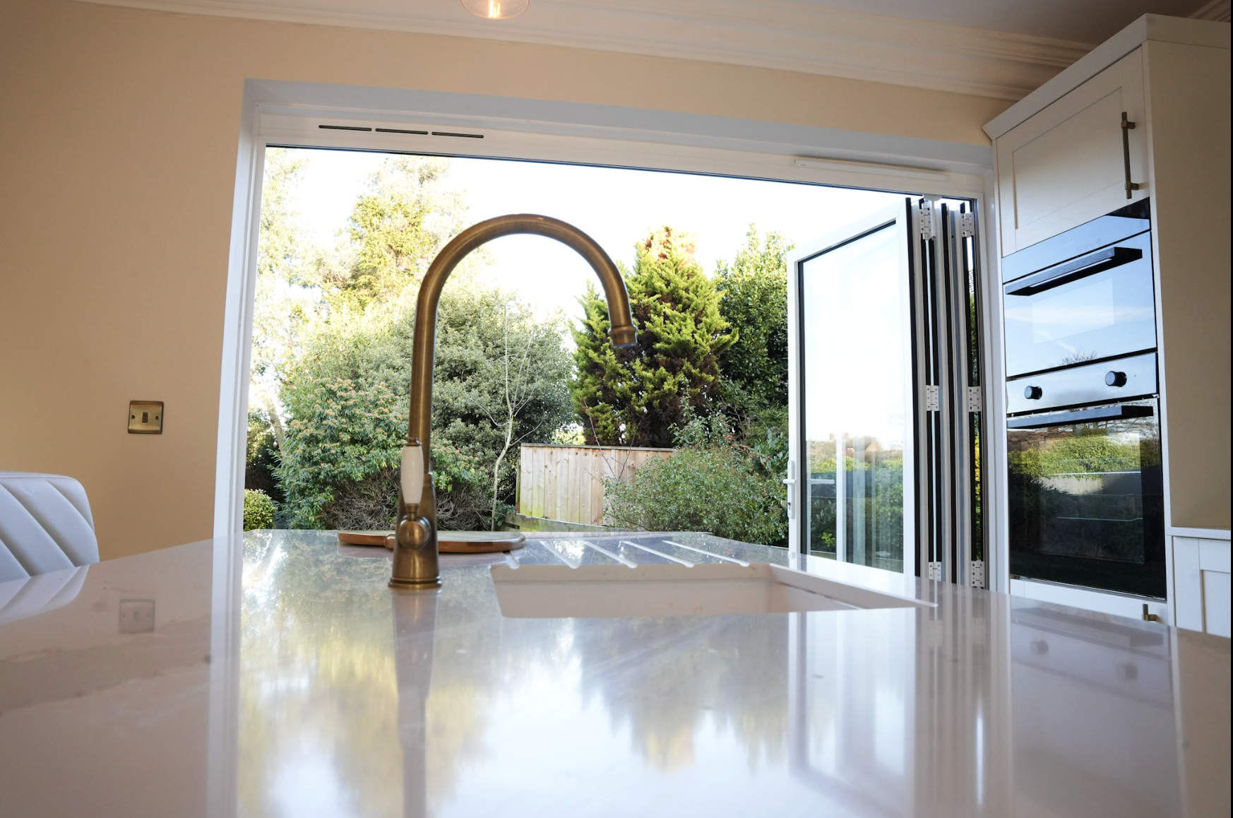 View of a bright kitchen with a large open window showing green trees outside, a gold-colored kitchen faucet, and cabinetry with an oven and microwave.